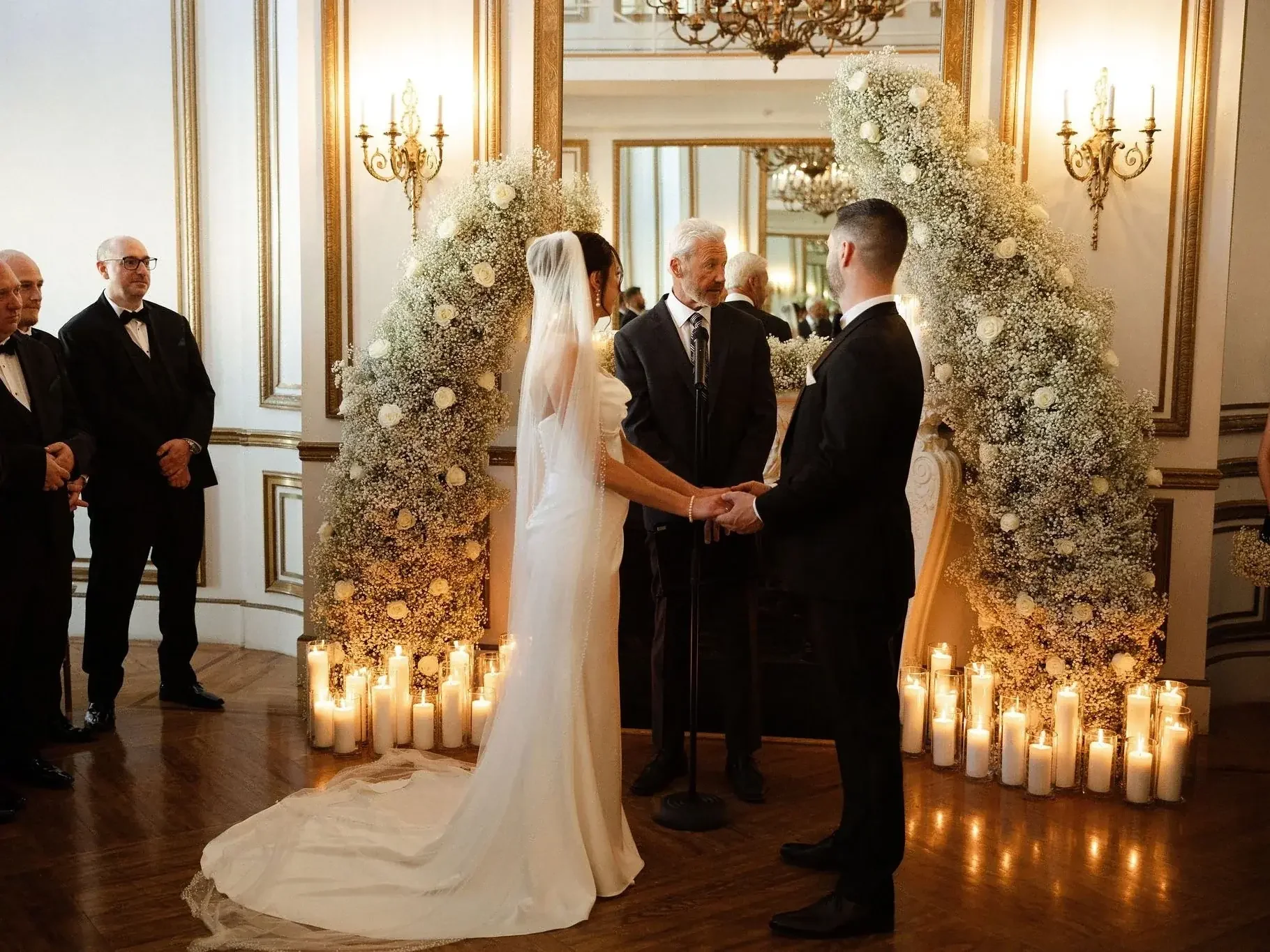 A wedding ceremony with a bride and groom holding hands in front of officiant, surrounded by floral arrangements and candles, with guests observing in an elegant, chandelier-lit room.