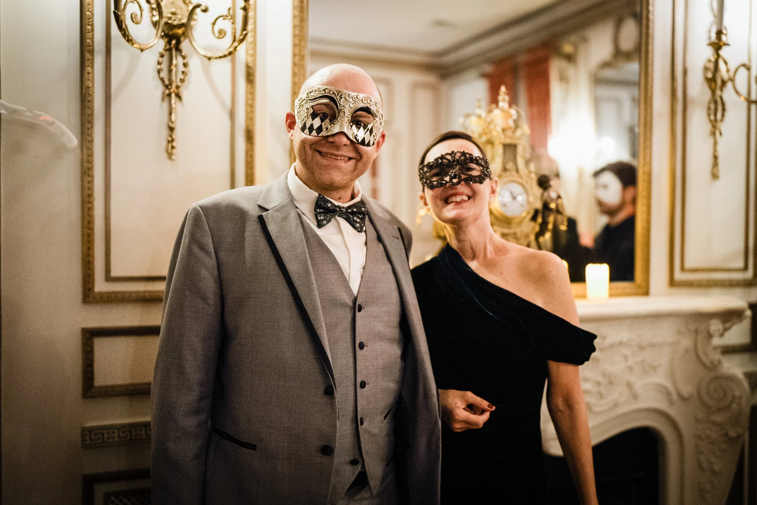 Two people at a fancy event wearing masks and formal attire, with ornate mirrors and decorative gold accents in the background.