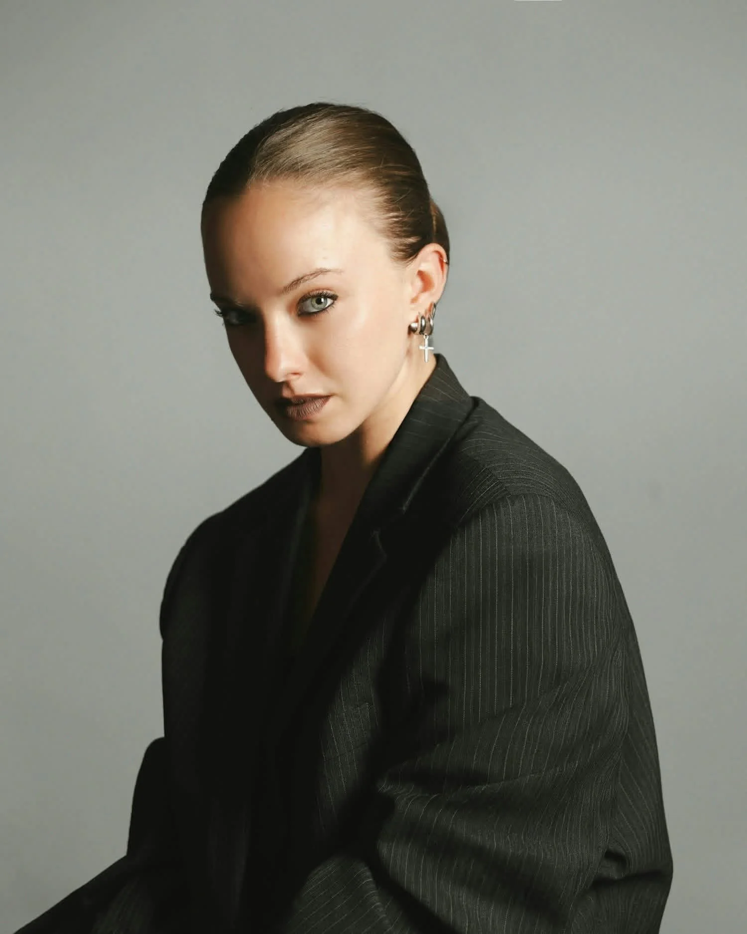 Glenda Gheller, Dance Artist & Choreographer. A woman with slicked-back blonde hair, wearing a black pinstripe blazer and cross earrings, looking at the camera with a neutral expression against a plain gray background.