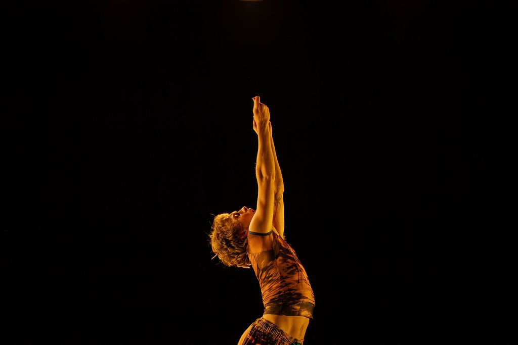 A dancer performing a graceful pose with arms raised overhead against a black background.