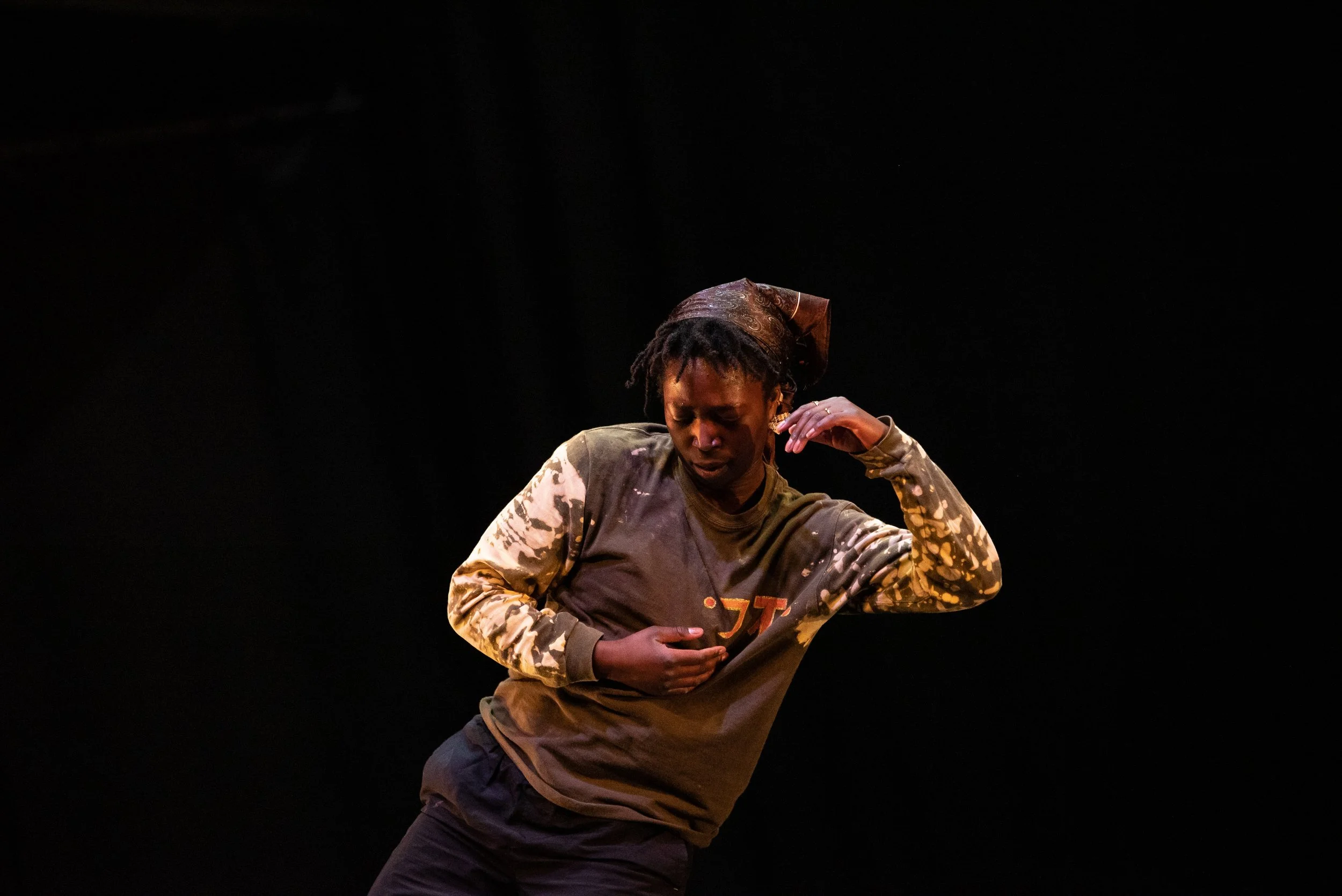 A young woman with dreadlocks is performing on stage, wearing a gray and beige sweatshirt with a patterned sleeve and a headscarf, with a dark background.