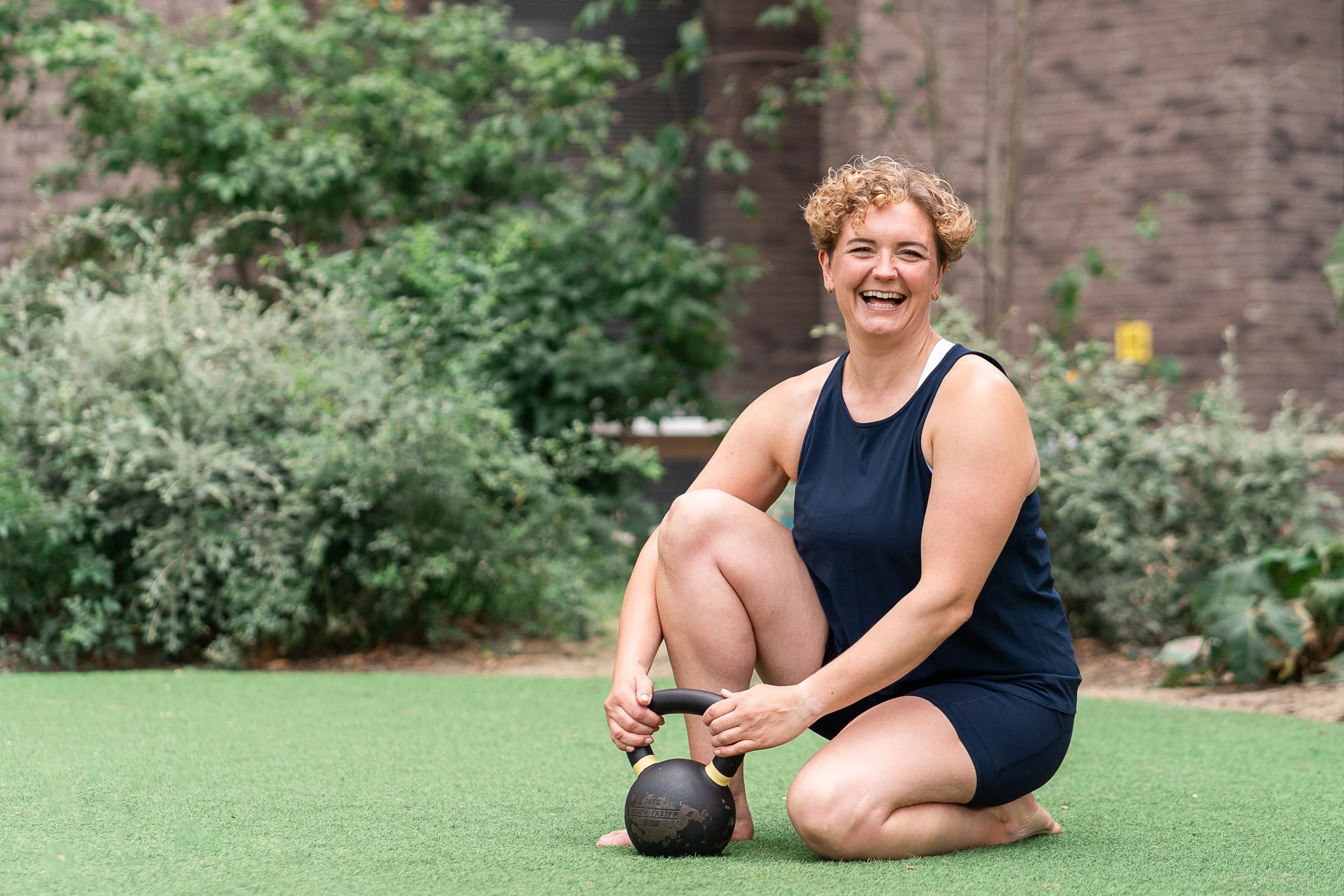 Strength coach Rowena Ramsell laughing while sitting on the grass beside a kettlebell