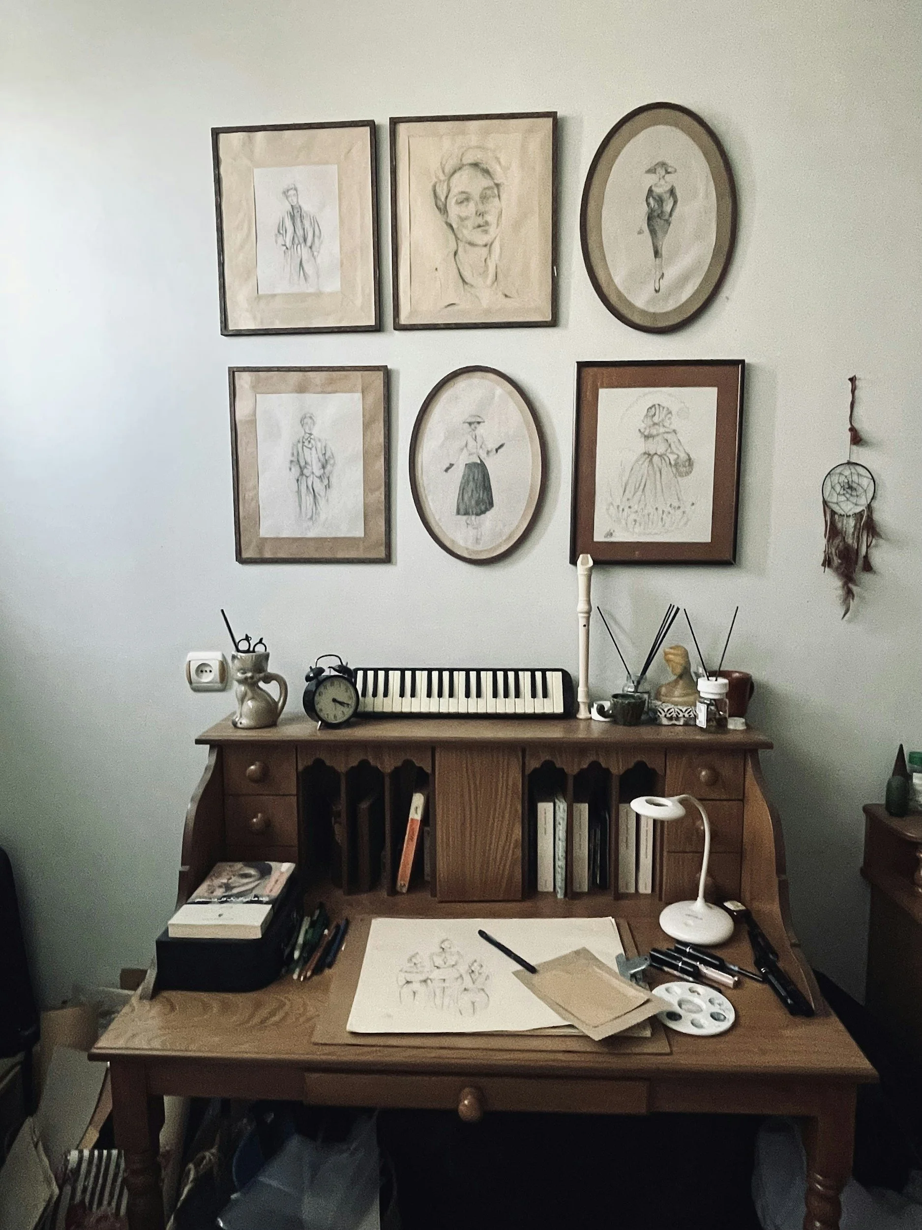 A wooden desk with drawing supplies, a lamp, books, and sketch papers, with framed artwork and a dreamcatcher hanging on the wall behind.