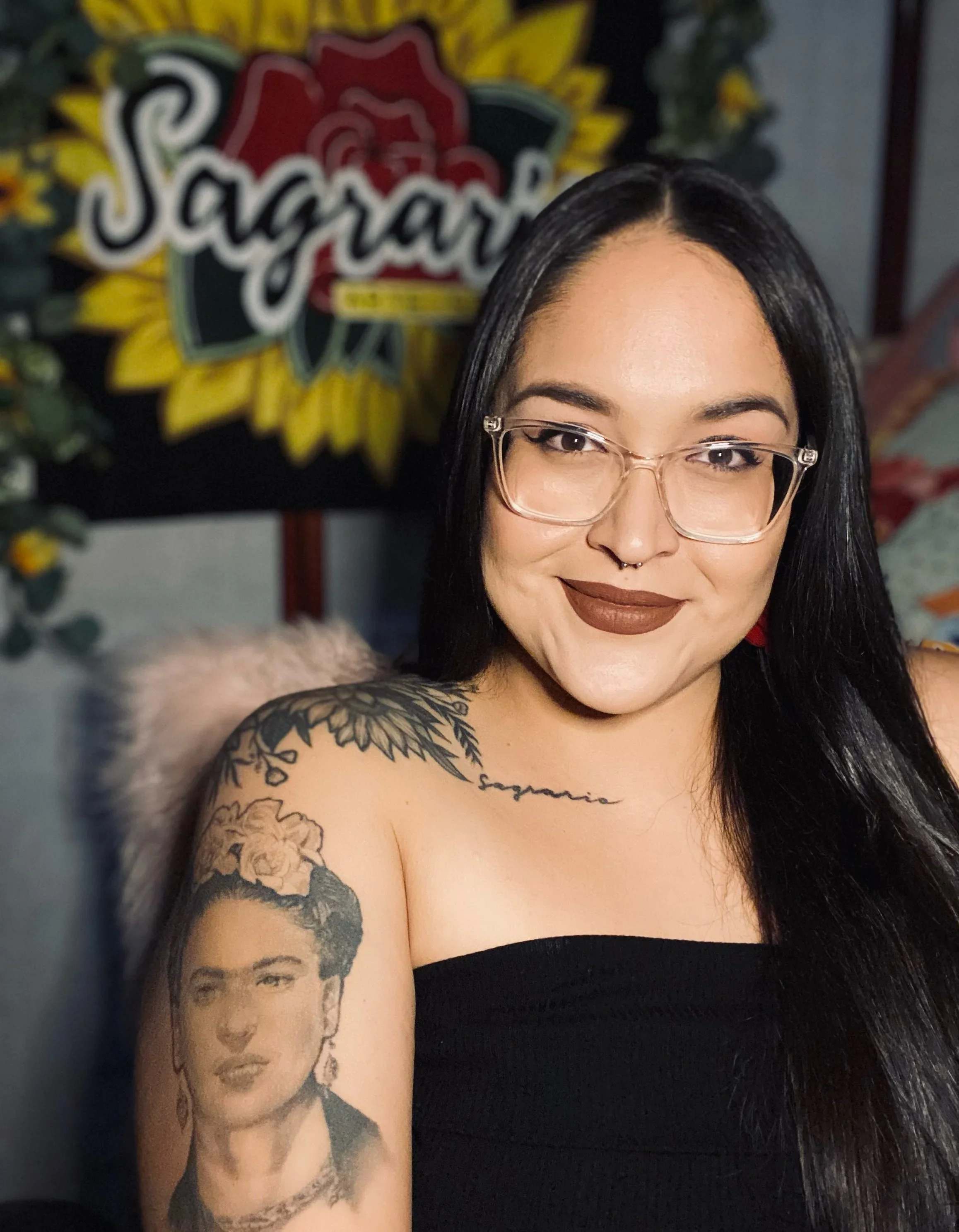 A woman with long black hair, glasses, and brown lipstick, wearing a black strapless top, showing tattoos of Frida Kahlo on her arm and a script tattoo on her collarbone, smiling in front of a sunflowers and rose sign that says 'Sagrado'.