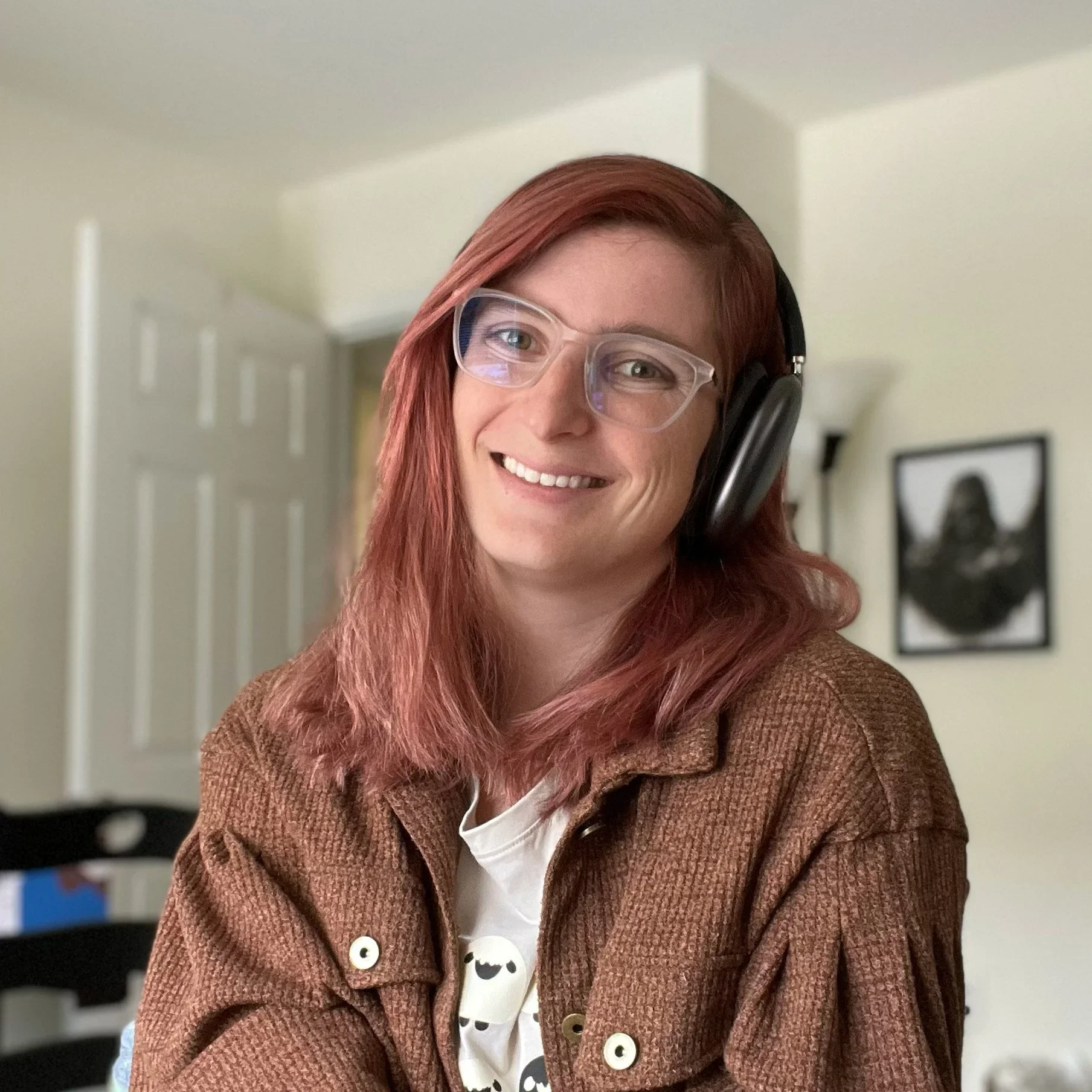 A portrait of Emma who has pink auburn hair, clear glasses, and is wearing a brown button up coat with a cream graphic tea. She's smiling and looks incredibly friendly and end in Dearing.