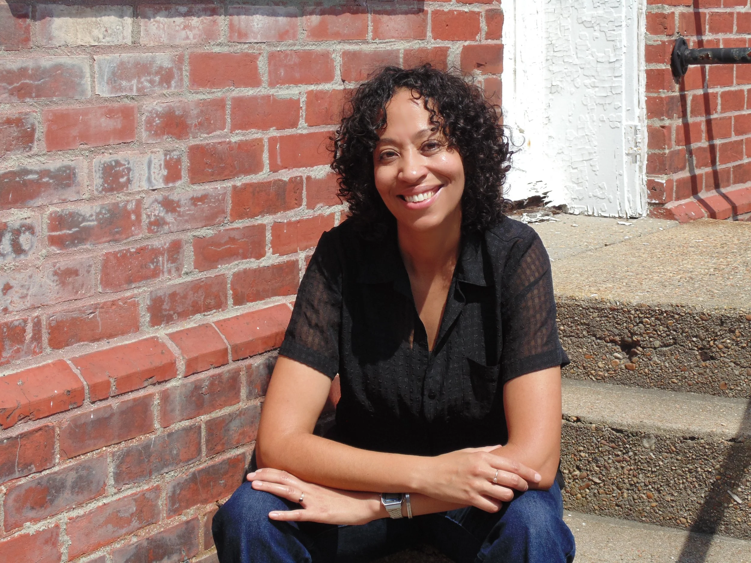 Adrienne G. Perry sits on smiling on concrete steps of a vacant brick building in Fort Monroe.