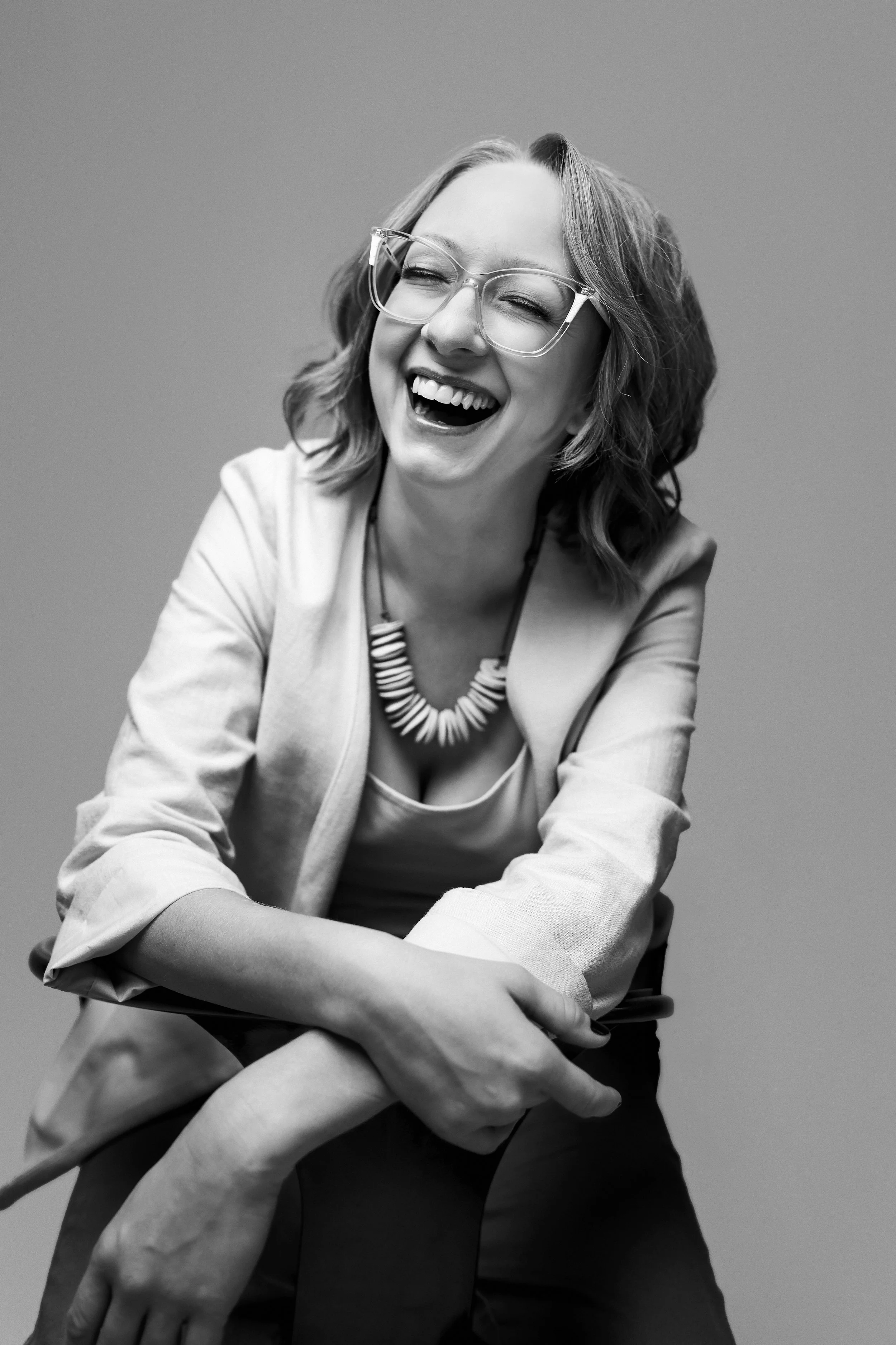 woman with short blonde hair and glasses leans up against a hair and laughs during her headshot session at opya studios in pittsburgh