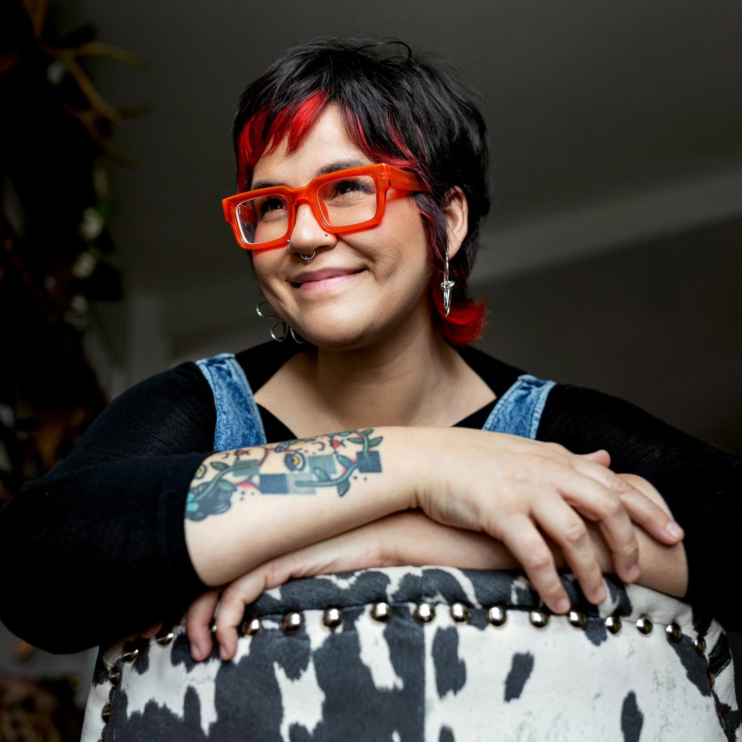 pittsburgh pa photographer tori stipcak leans over a cow print chair. she is wearing bright red glasses, has hort brown hair, and is wearing overalls