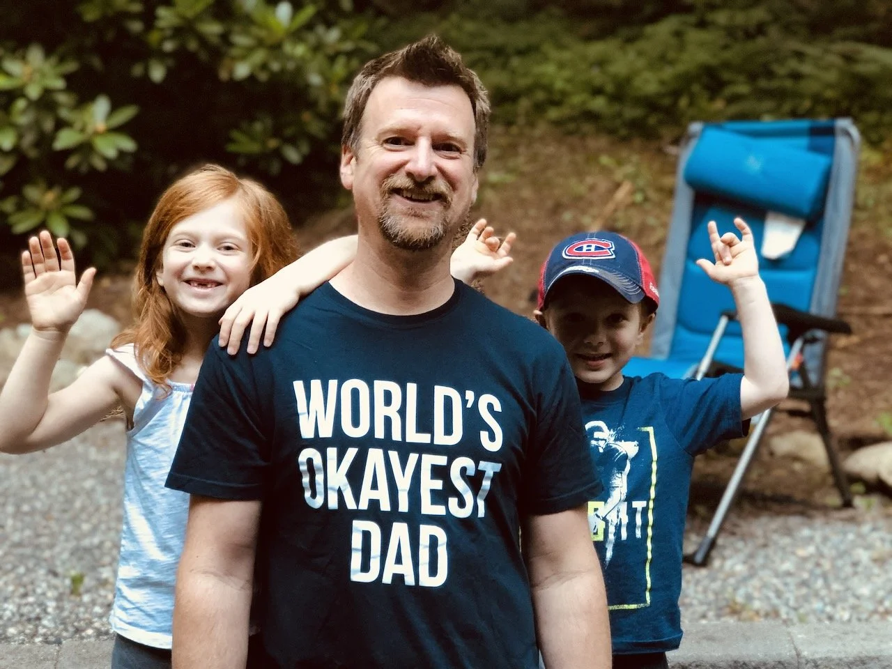 A smiling man with two children behind him outdoors, with a blue folding chair in the background. The man wears a black T-shirt with the text 'WORLD'S OKAYEST DAD'. The girl has red hair and waves, the boy wears a baseball cap and a T-shirt, and they appear to be happy.