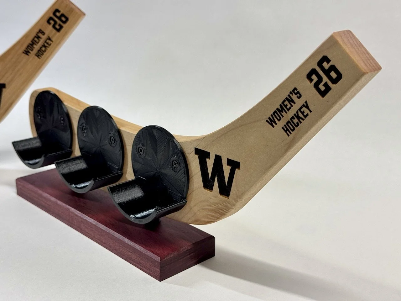 A wooden hockey stick with a black 'W' on the blade, labeled 'Women's Hockey 26,' positioned on a red base, with three black hockey pucks in front.