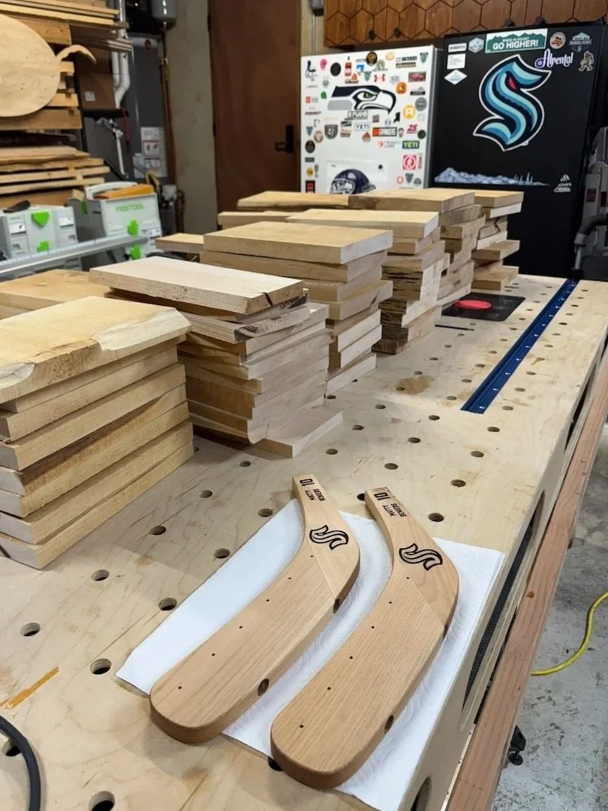 Two finished shoehorns made of light-colored wood on a workbench in a woodworking shop, surrounded by stacked uncut wood pieces and woodworking equipment.