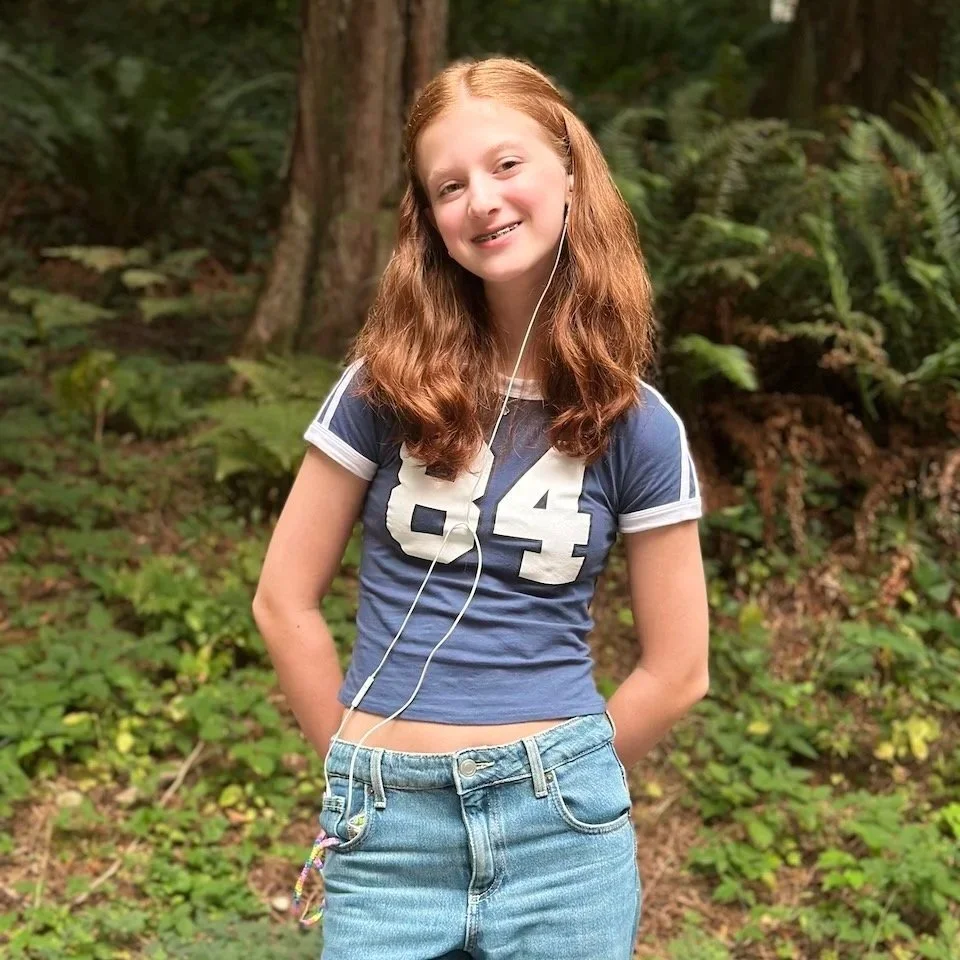 A young girl with red hair smiling outdoors in a wooded area, wearing a navy blue crop top with the number 84 on it and light blue jeans, with earphones in her ears.