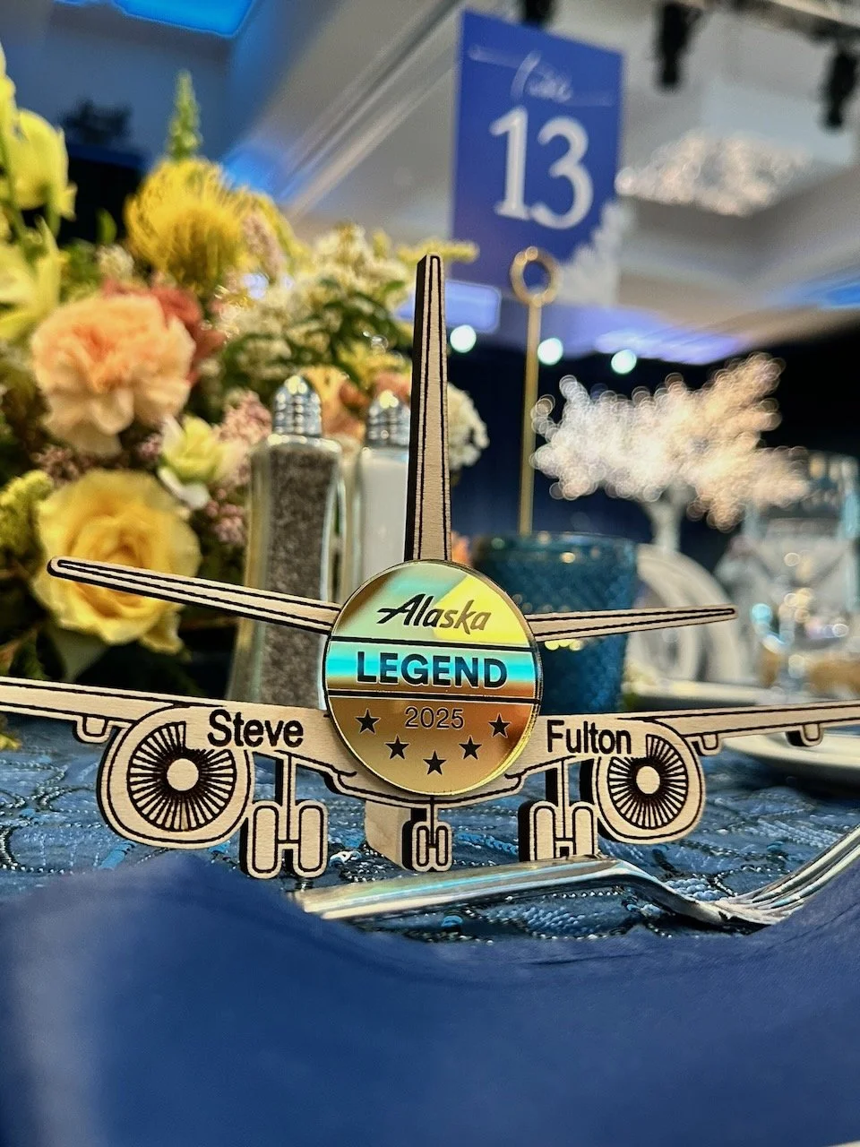 A decorative airplane-shaped award with the words "Alaska Legend 2025" and stars in the center, placed on a table with a blue tablecloth. In the background, there are colorful flowers, condiments, and a table number 13 sign.