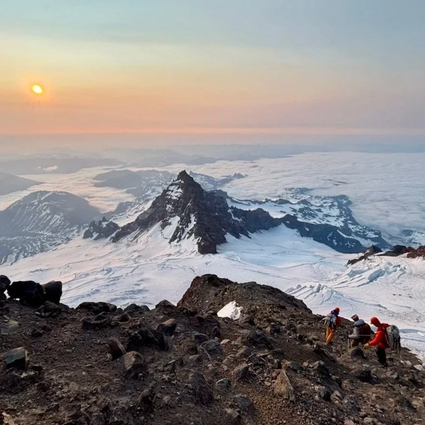 Mountaineers climbing a rocky slope with a snowy mountain and glacier in the background, under a sunset sky.