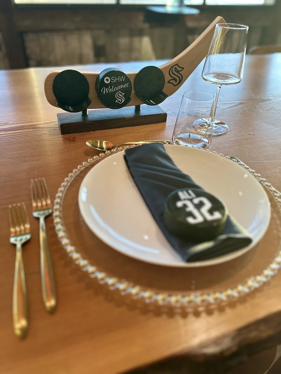 A dinner table setting with a white plate, black napkin, gold utensils, two water glasses, a wine glass, and a decorative table marker with a hockey puck that reads '32' and the inscription 'ALL TIME.' A wooden playful hockey stick-shaped decor with 