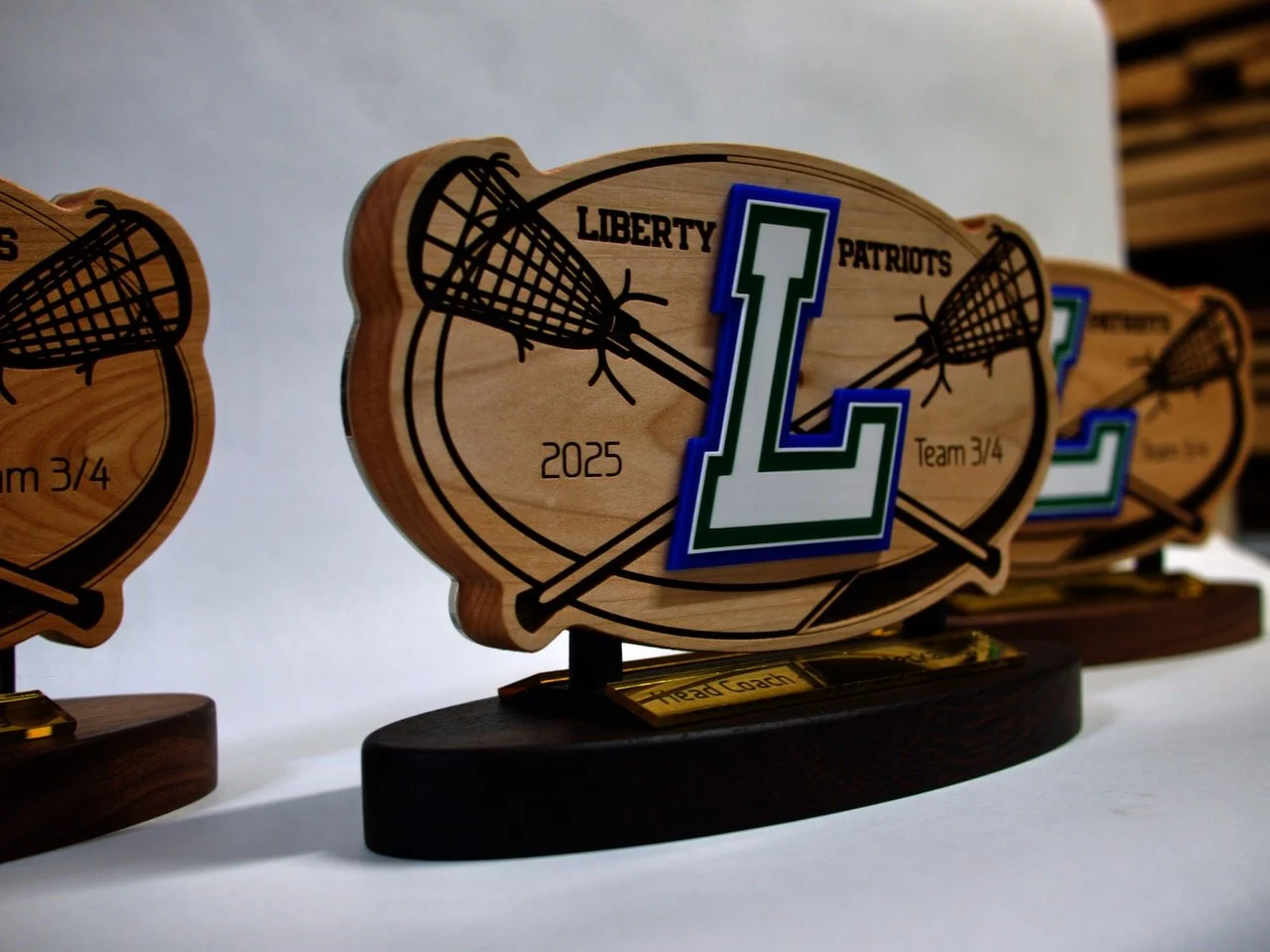 Wooden trophy with a lacrosse stick graphic, labeled 'Liberty 2025,' featuring a large green and blue letter 'L' in the foreground, on a dark wooden base.