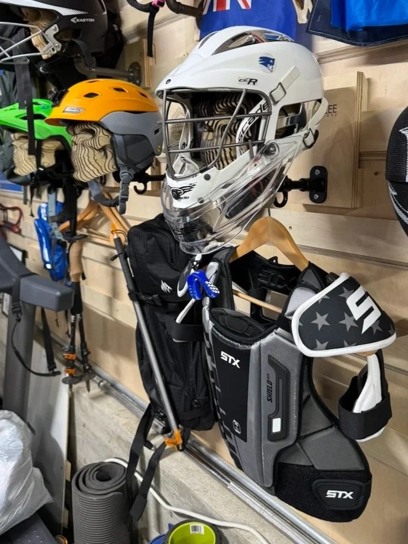 Lacrosse and snowboard helmets, including a white helmet with a clear visor, a yellow and gray helmet, and a black helmet, displayed on a wall in a store, with other riding gear and accessories visible.