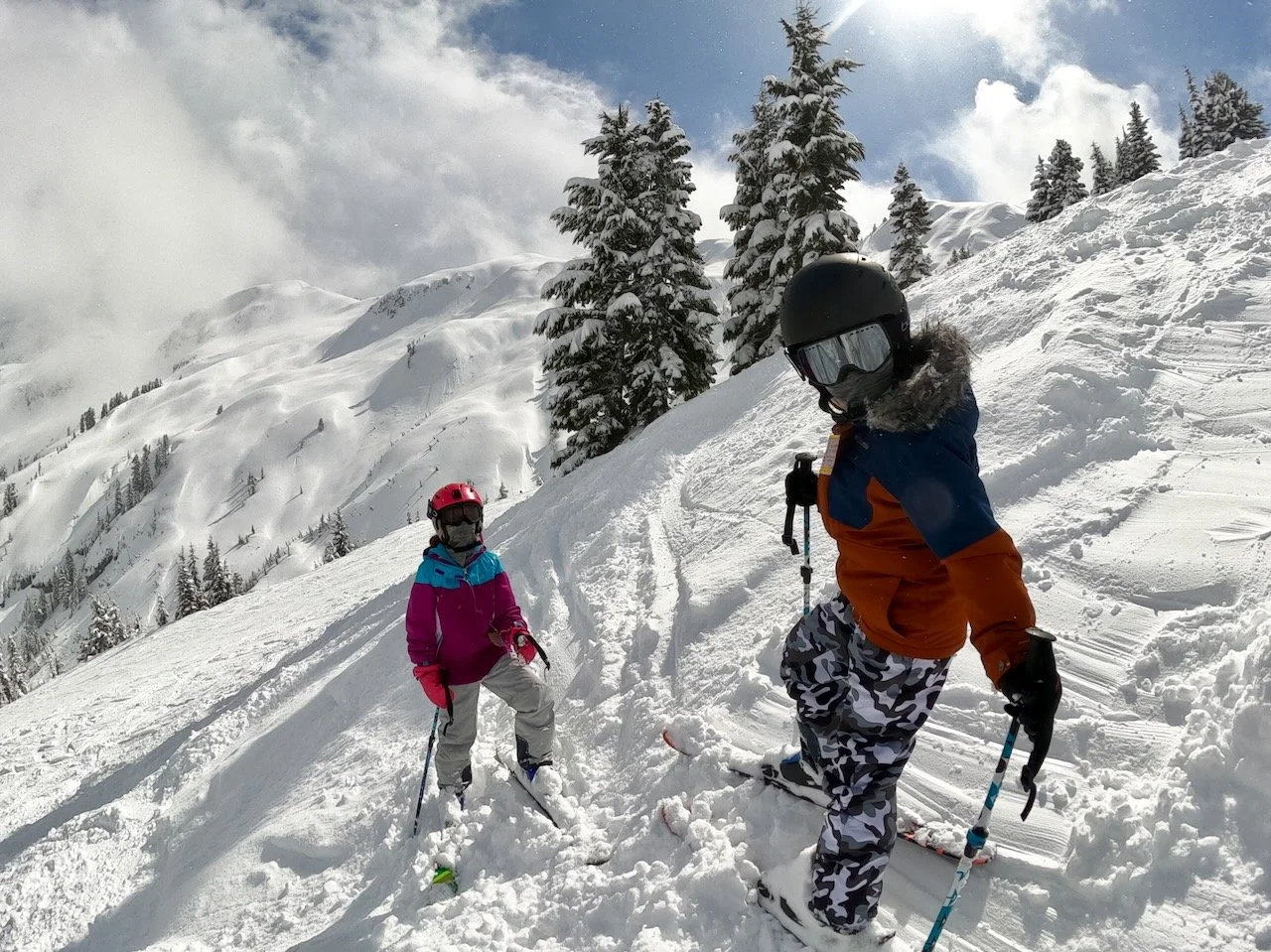 Two children in colorful winter clothing and helmets skiing on a snowy mountain slope with snow-covered trees and mountains in the background.