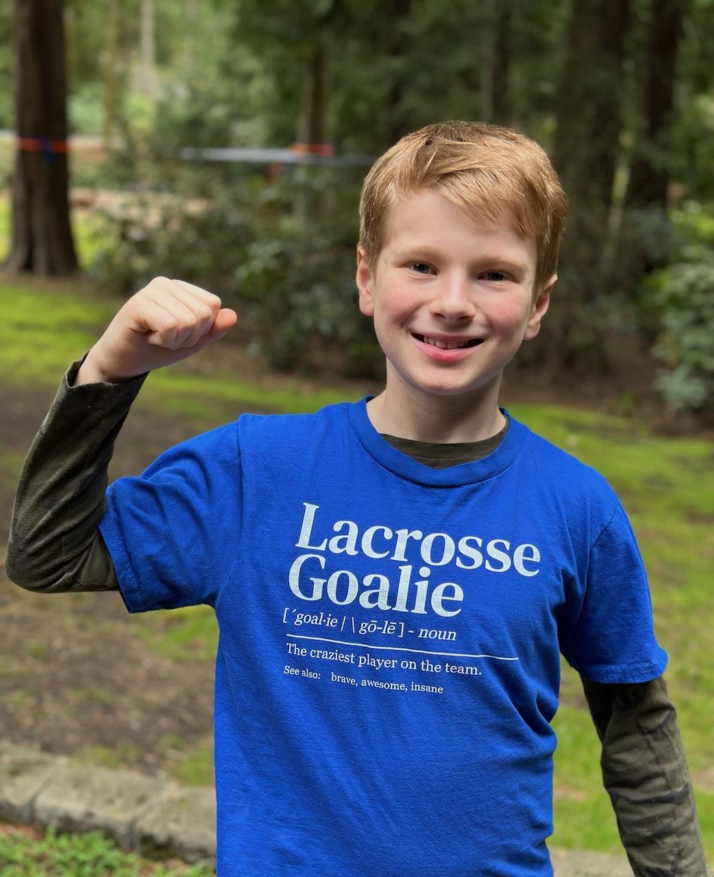 A young boy with red hair smiling and flexing his right arm, outdoors in a wooded area, wearing a blue t-shirt that says 'Lacrosse Goalie' and includes a humorous definition.