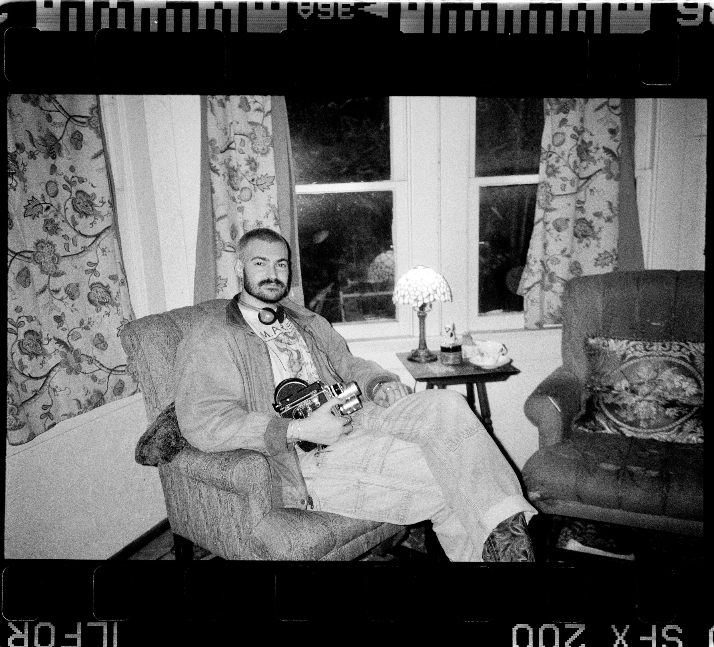 Black and white photo of a man sitting indoors with patterned curtains behind him. He has a beard and is holding a camera. There is a small lamp on a table beside him.