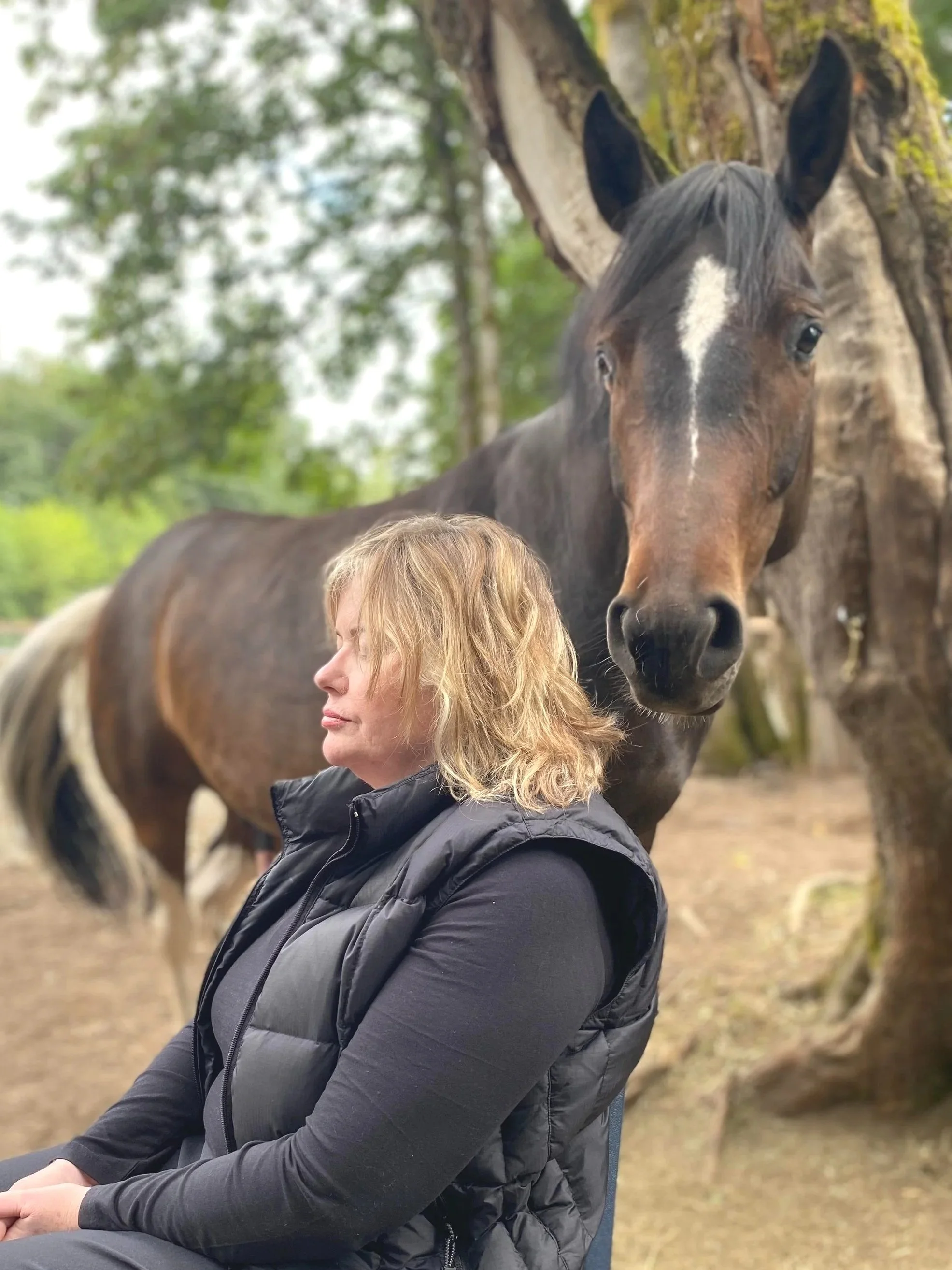 A woman with blonde hair wearing a black jacket sitting outdoors near a dark brown horse with a white star-shaped mark on its forehead. The background features trees and a cloudy sky.