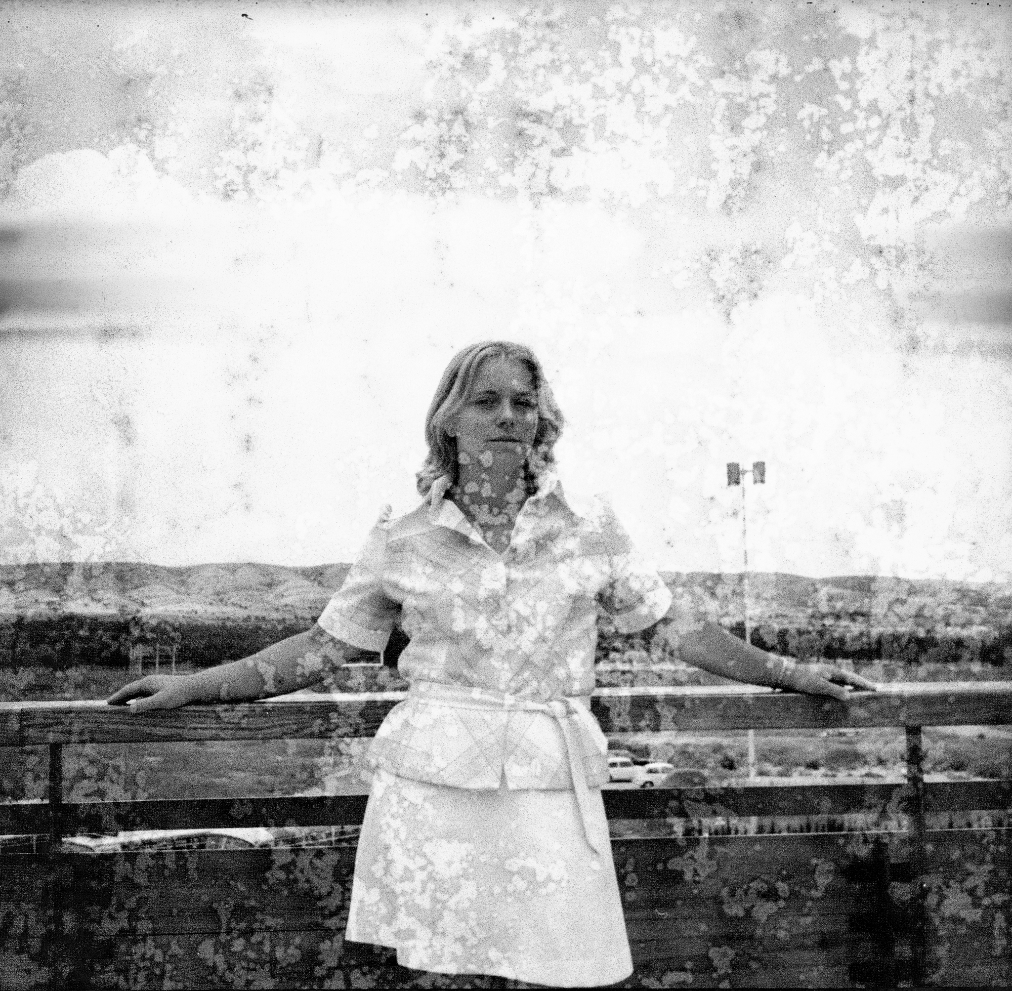 Image Description: In this image we have a teen or young adult stood in front of some fencing which is the same as the images with two young children (boy and girl) which are placed after photograph #3. The teen/young adult is leaning on the fencing, both her arms on either side placed on the top plank of the fence. She is wearing a white skirt, a light coloured plaid shirt with sleeves that end just before the elbow. The young lady has shoulder length light coloured hair. Not much scene in this photograph, except for sky, some green-land, a car park and the fencing behind her.