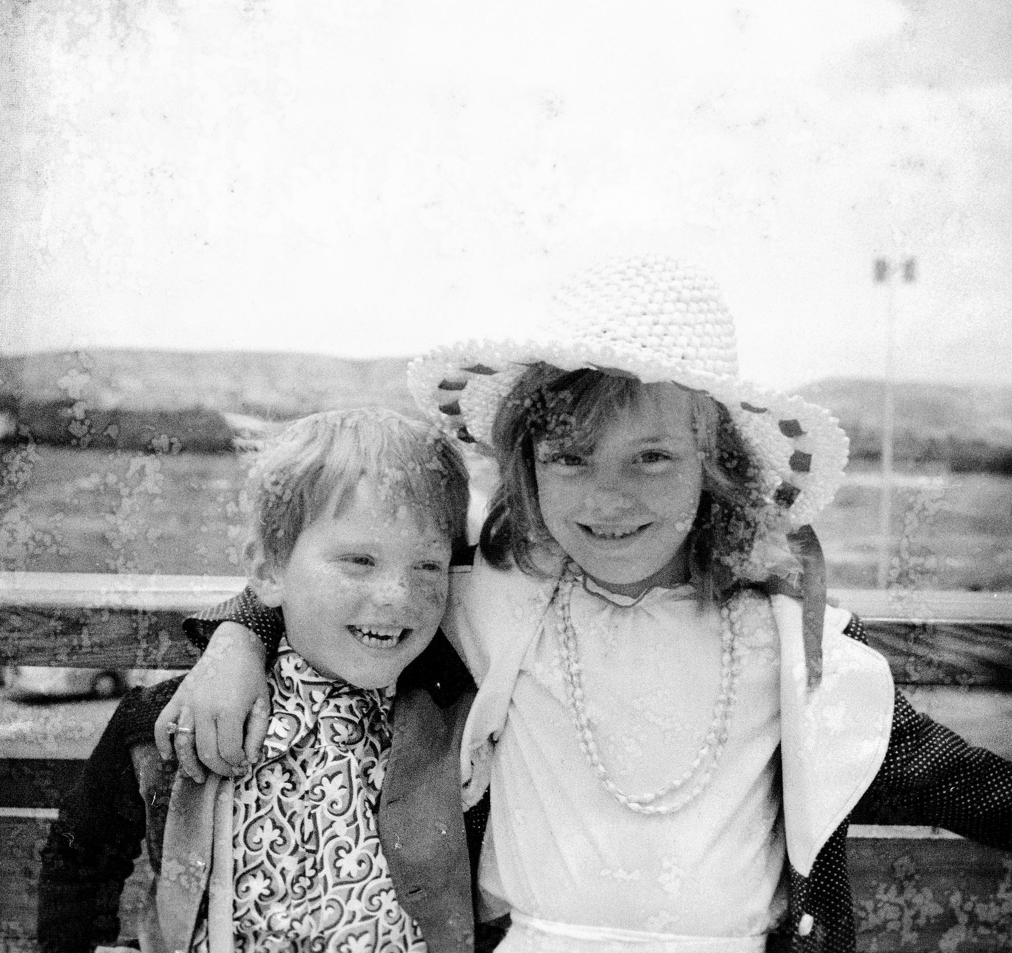 Image Description: Photograph features a young boy and a girl. The girl (right) is wearing sun hat, her hair down, with a white dress, what seems to be pearls around her neck, and a white and grey/black jacket. Polka-dots are on the sleeves and body suit of the coat. The boy (left) is wearing a black and grey jacket, a patterned scarf with trousers and white suspenders that can’t be clearly seen the photograph. The girl is looking at the camera smiling, the boy is looking to our right, also smiling. The girl has her right arm around the boy, a ring on the third finger, whilst her left hand trails of camera, leaning on the wood plank that goes across the image. Not much scene in this photograph, except for sky, some green-land, a car park and fencing behind them, which they seem to be leaning on.