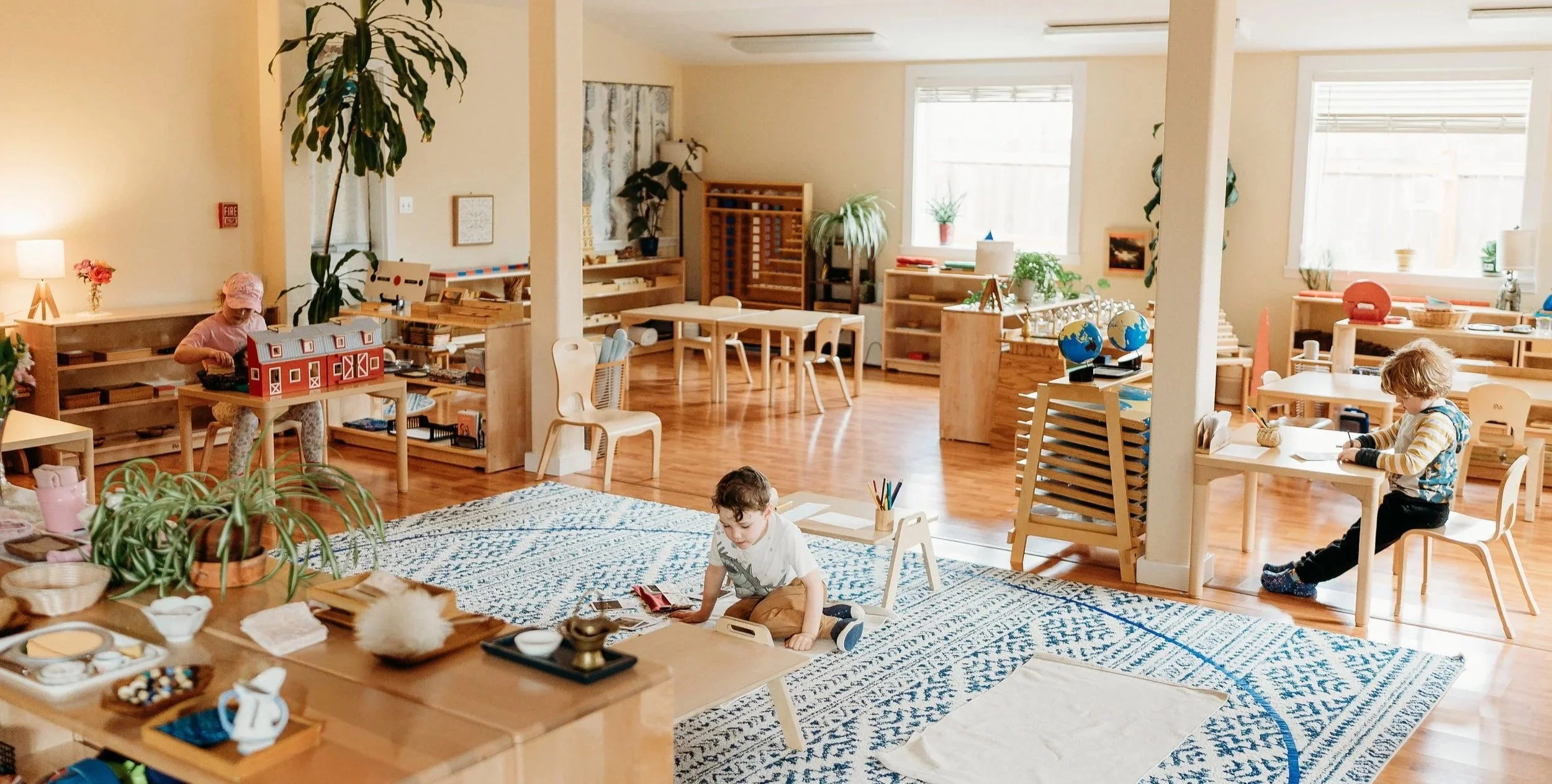 Children playing and learning in a bright, spacious classroom with wooden furniture, plants, and educational materials. Some children are reading, drawing, or playing on the floor.
