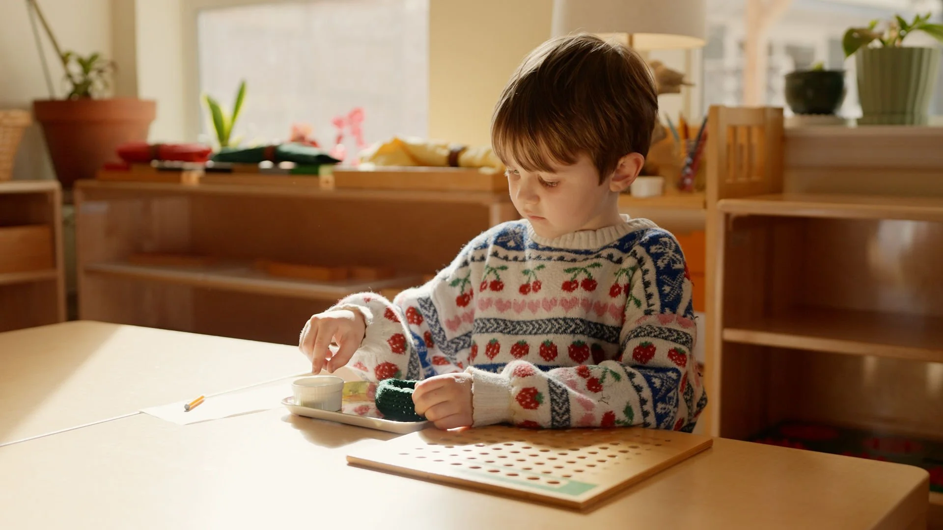 A young boy sitting at a table, playing a board game with a small cup and some game pieces, wearing a Christmas sweater with a cherry pattern inside a cozy room with wooden furniture and potted plants.