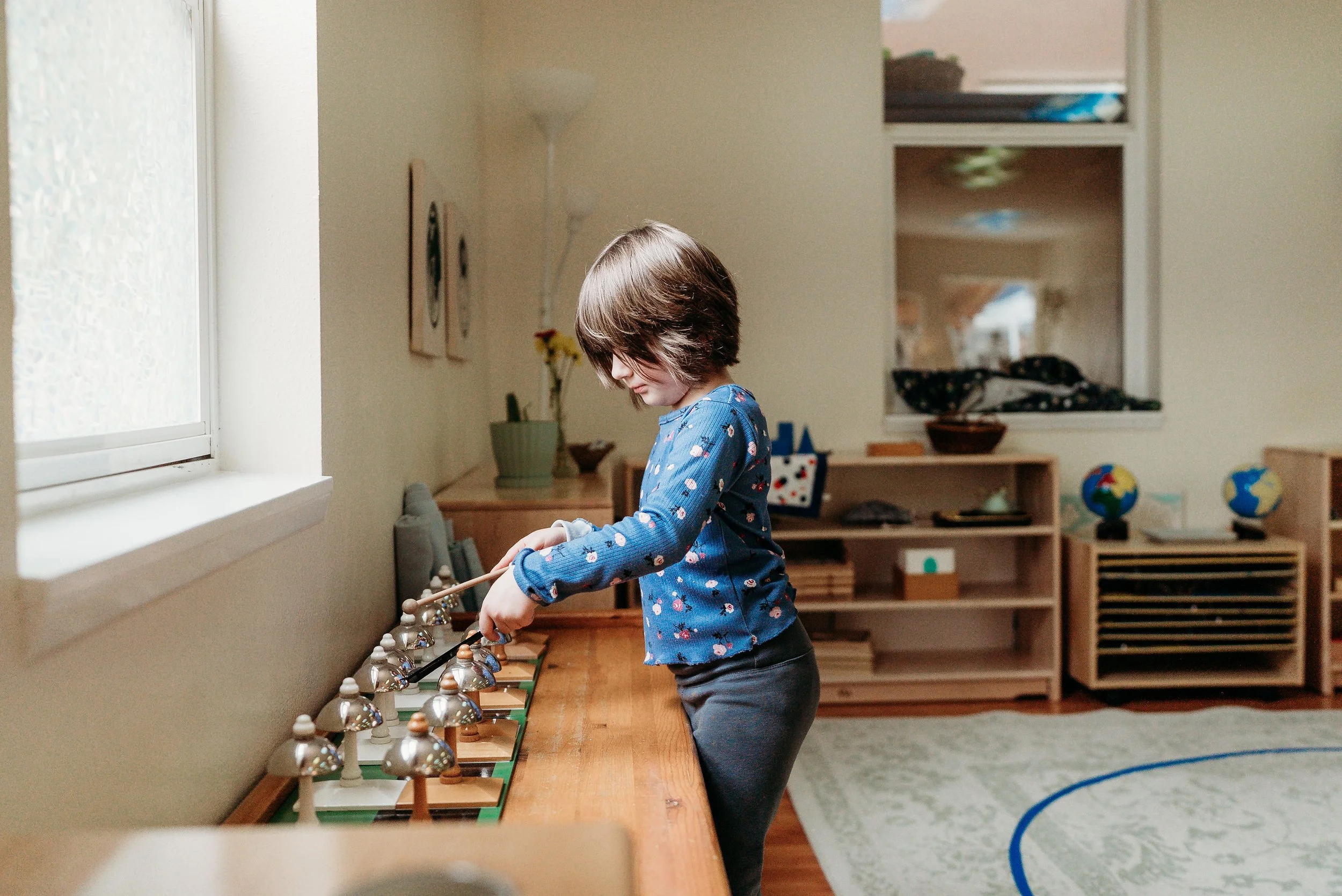 A young girl with brown hair in a blue shirt with colorful patterns playing chess at a wooden table in a brightly lit room.