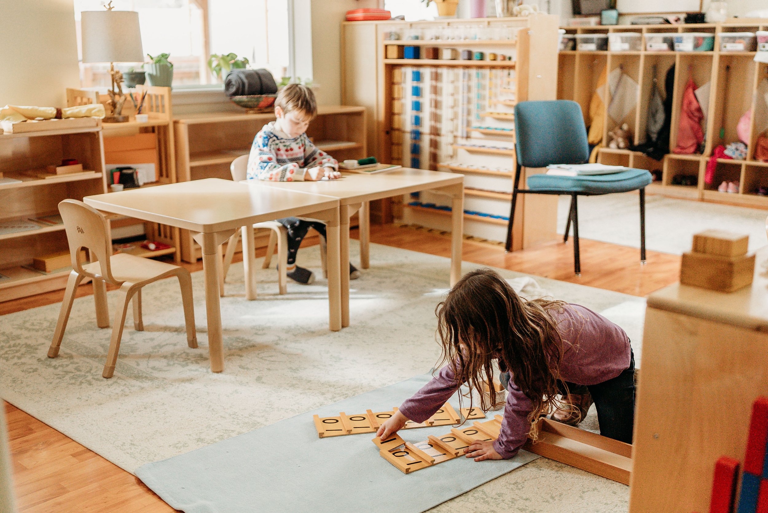 A young girl in a purple shirt and black pants playing with wooden educational tiles on a soft mat in a classroom. The classroom has light wooden furniture, shelves filled with books, toys, and supplies, and large windows letting in natural light.