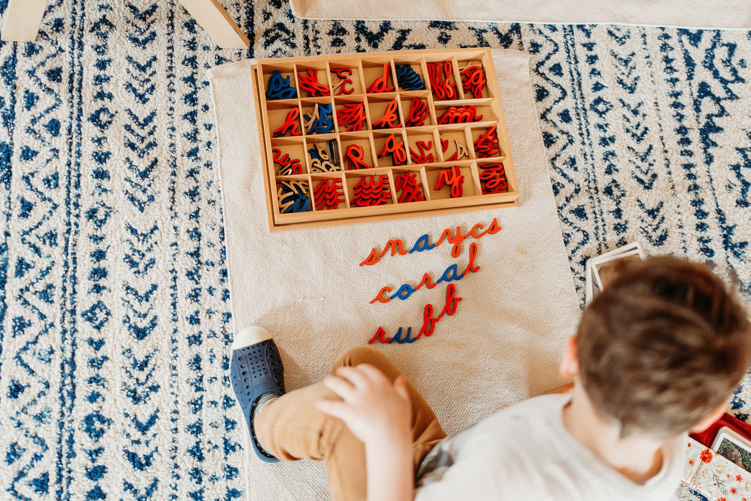 Children playing a game with wooden letter tiles on a beige cloth, with a blue and white patterned rug underneath.