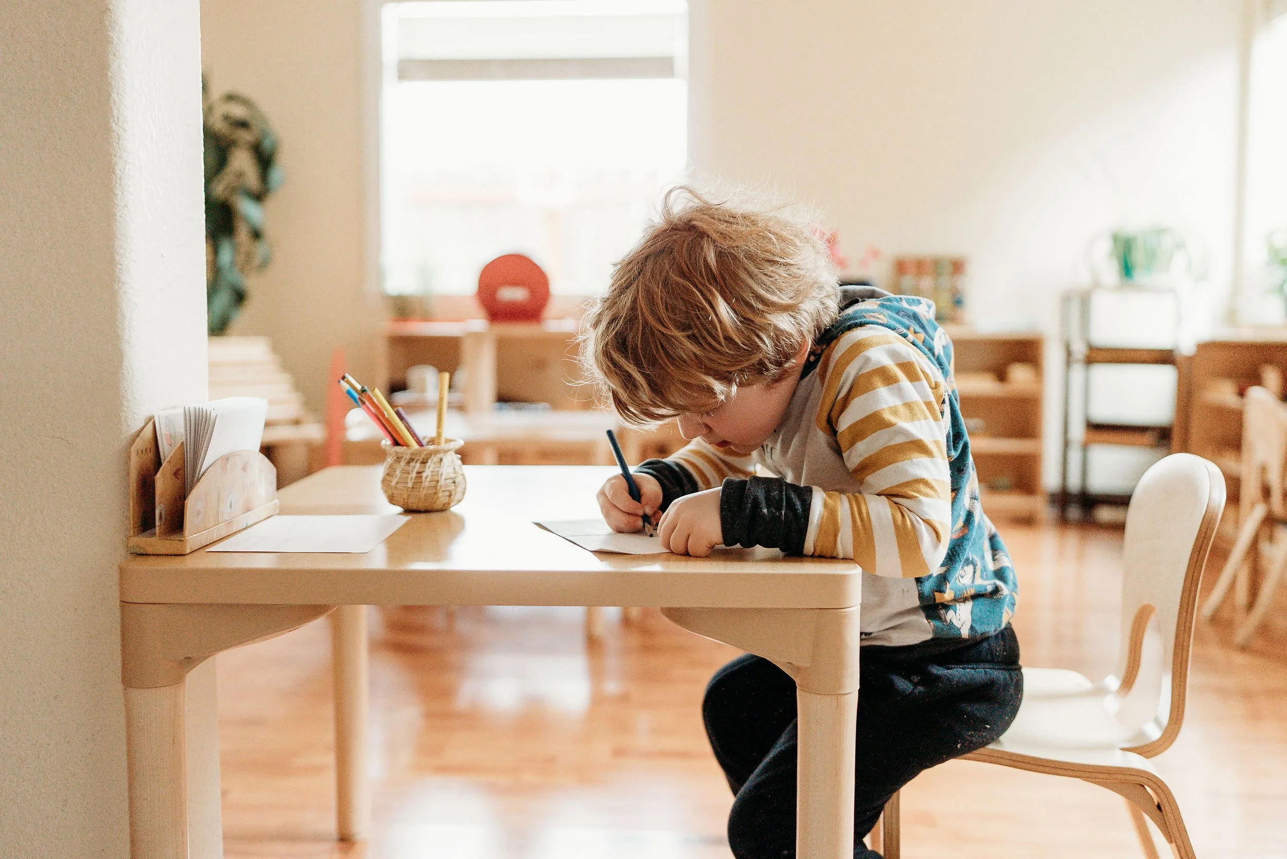 A young child with light brown hair, wearing glasses, a striped long sleeve shirt, and a hoodie, sitting at a small desk and writing on paper with a pen in a brightly lit classroom.