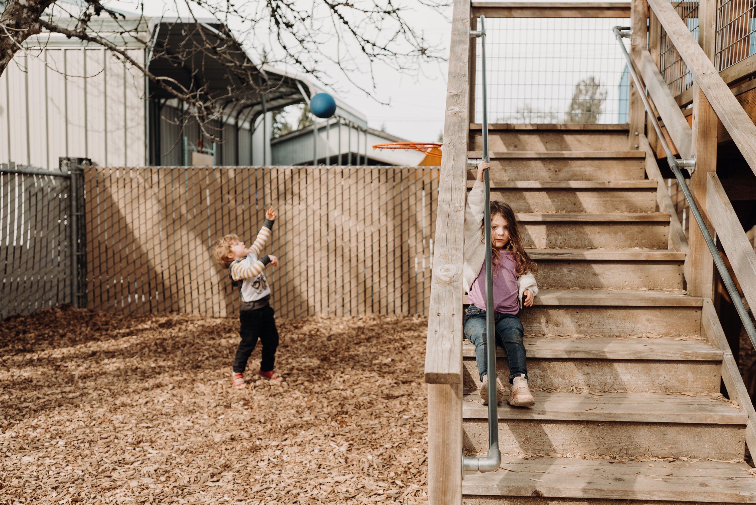 Two children playing outdoors near wooden stairs. One girl with long curly hair is sitting on the stairs, and a boy is reaching for a blue ball in the air. The area is fenced, with trees and a building visible in the background.