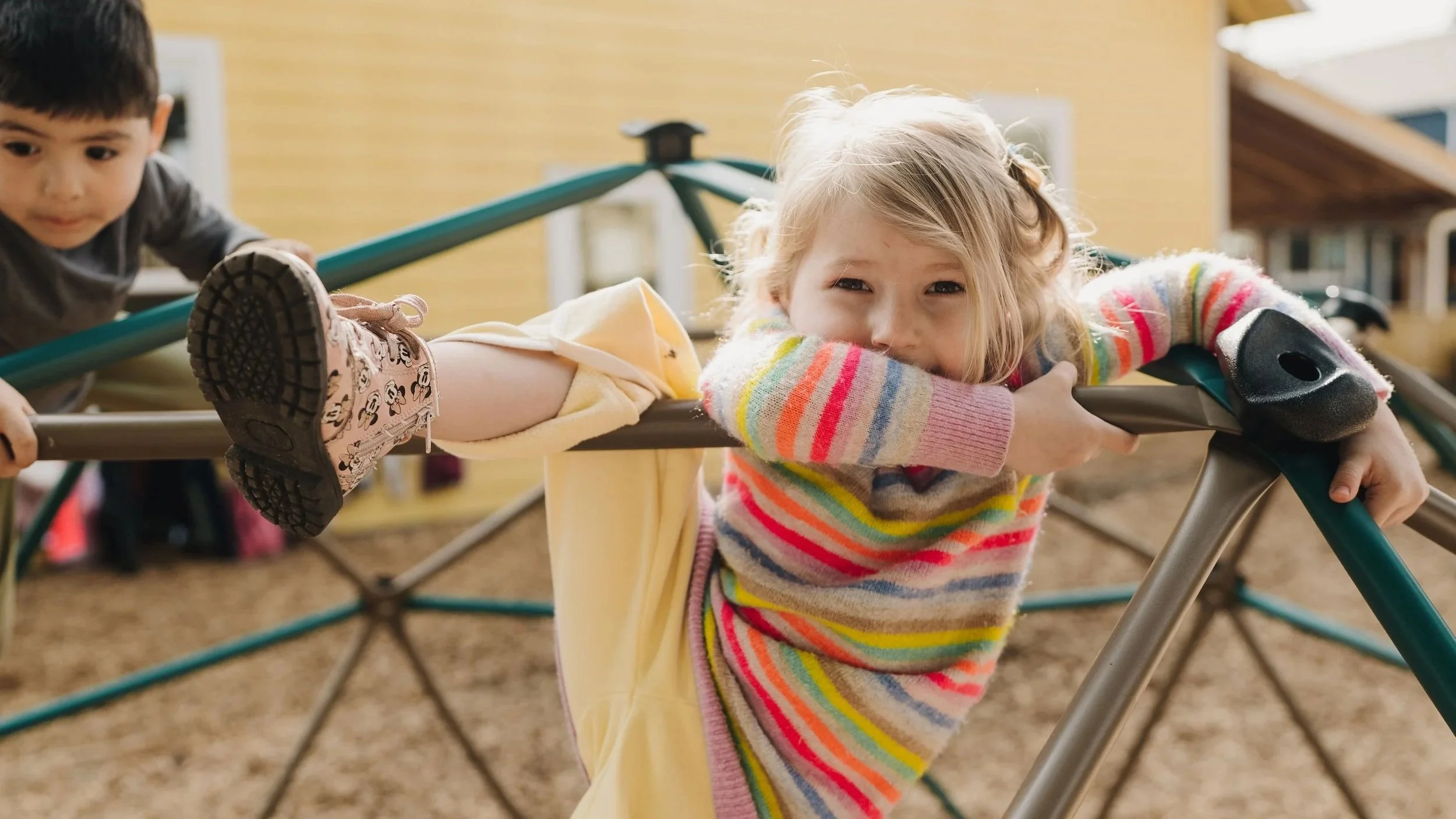 A young girl with blonde hair wearing a colorful striped sweater is lying on a playground slide, smiling with her head resting on her crossed arms at the top of the slide. A young boy with dark hair and a gray shirt is nearby, also on the playground.