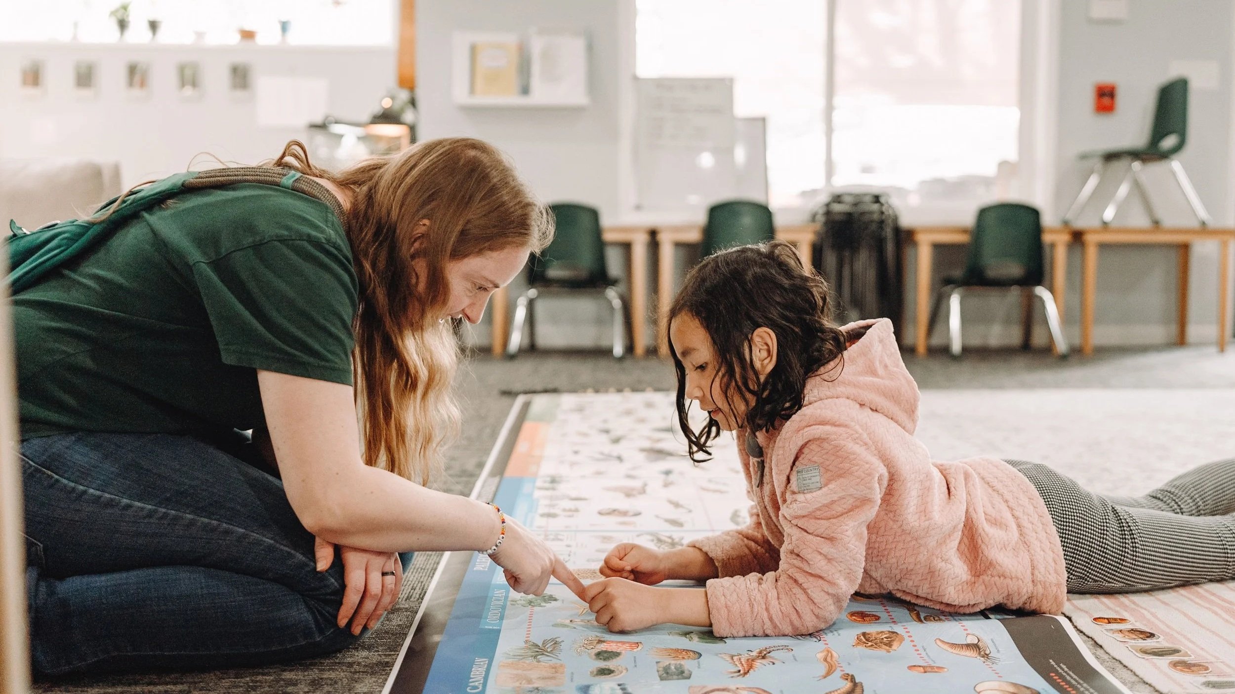 An adult and a young girl are lying on the floor, looking at a large illustrated poster with various animals and plants. The adult is pointing at the poster while the girl is observing attentively. The background shows a room with chairs, tables, and windows.