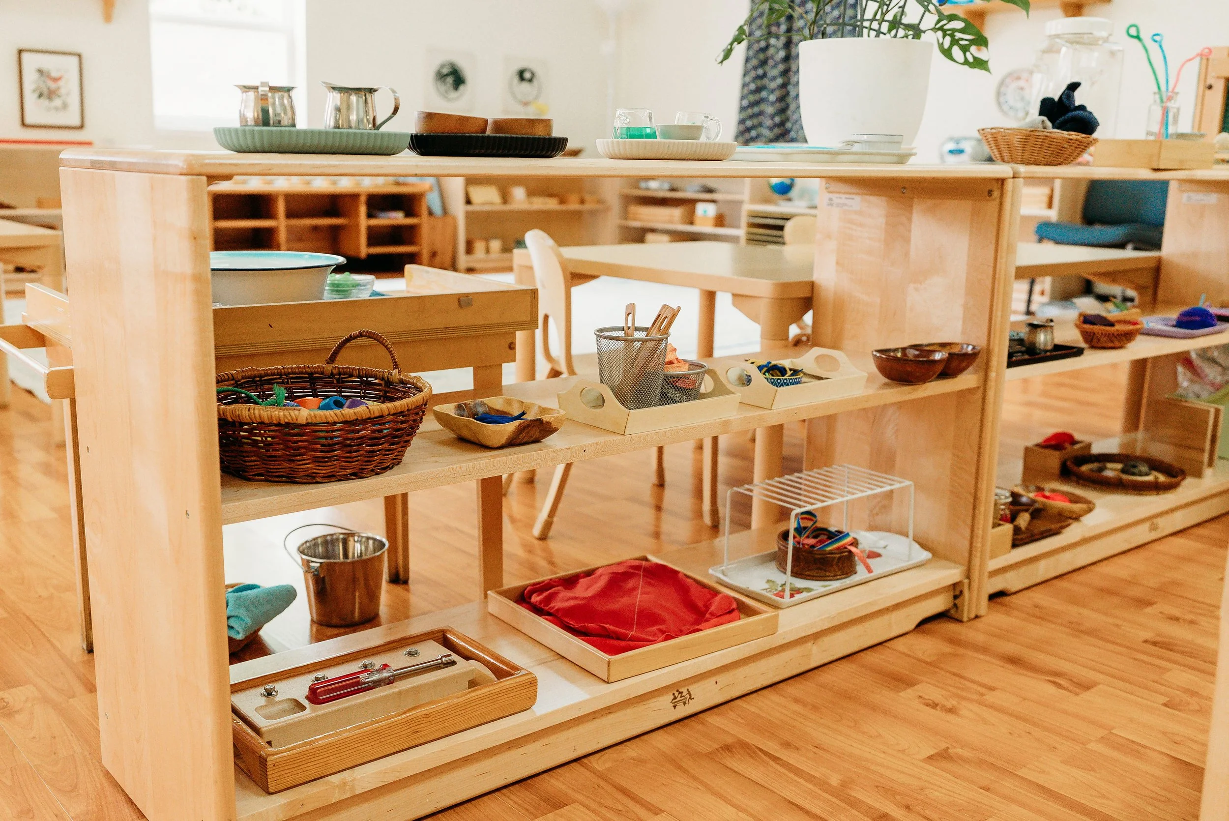 A wooden shelving unit with various children's toys and objects, including baskets, bowls, and containers, in a room with wooden floors and a background with additional shelves and decor.