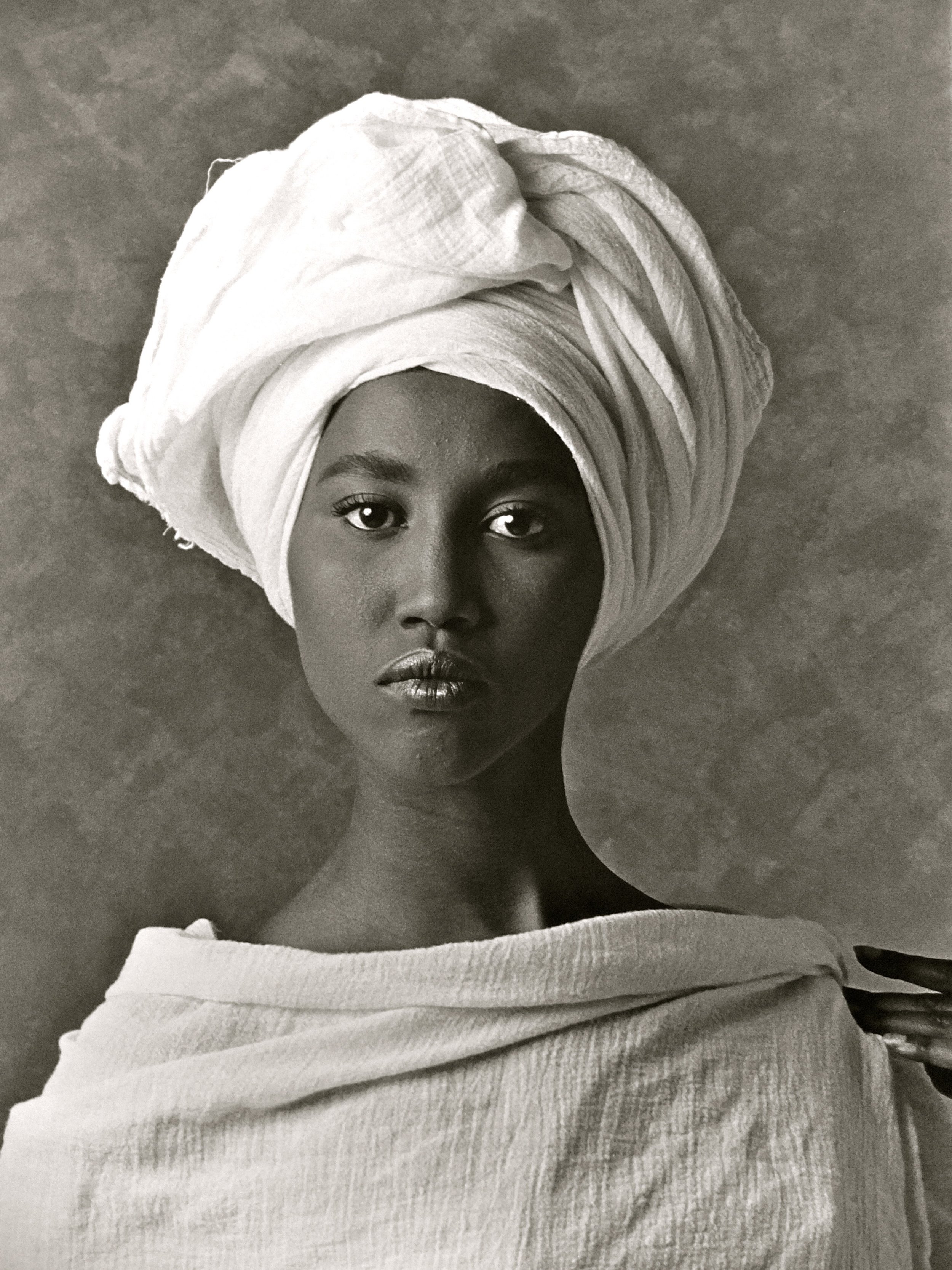  A black and white portrait of a woman of dark complexion from the clavicle up. She has a head wrap and garb made of the same white fabric. The fingers of her left hand rest on her left shoulder. 