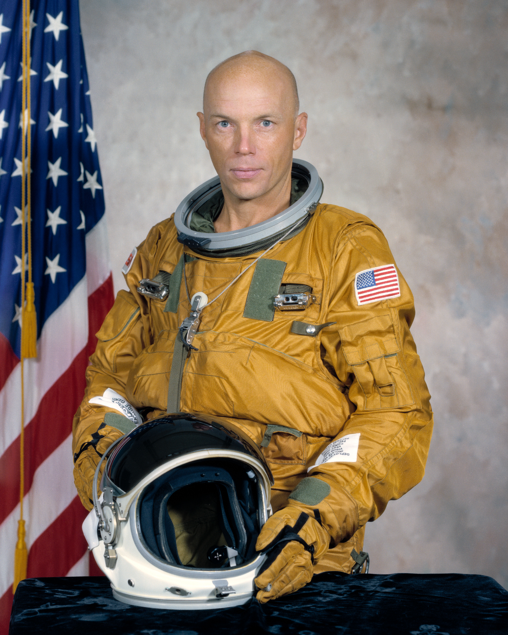 Story Musgrave wears a space suit beside an American flag.