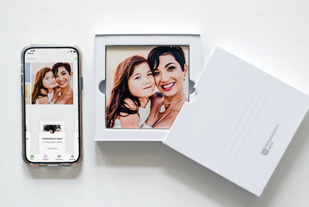 Persnickety Box photo printing app on a phone next to a box of square monthly photo prints