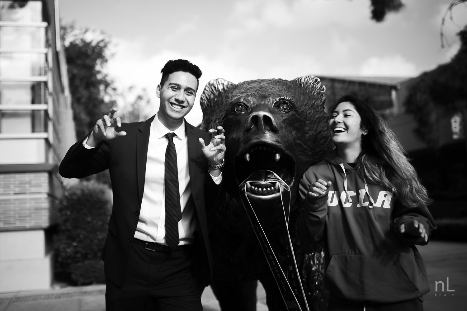 UCLA Class of 2019 graduate wearing suit and tie and graduation sash with girlfriend posing in front of Bruin Bear.