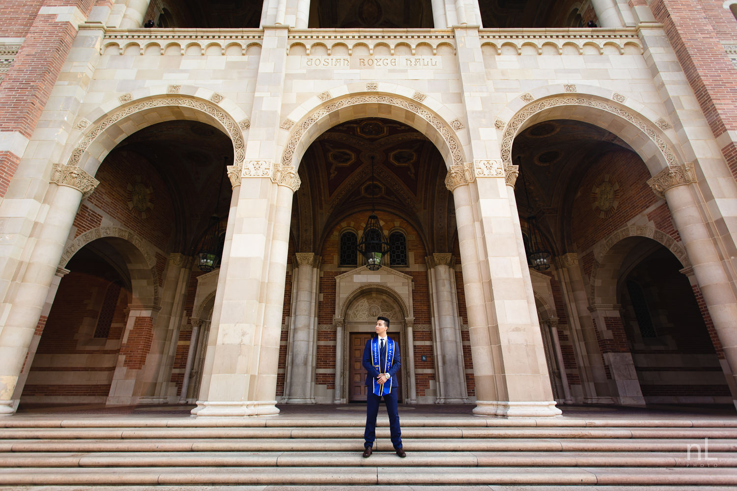 UCLA Class of 2019 graduate wearing suit and tie and graduation sash standing in front of Royce Hall.