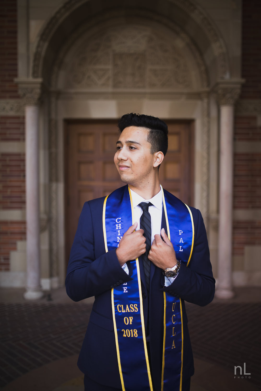 UCLA Class of 2019 graduate wearing suit and tie and graduation sash smiling and standing in front of Royce Hall.