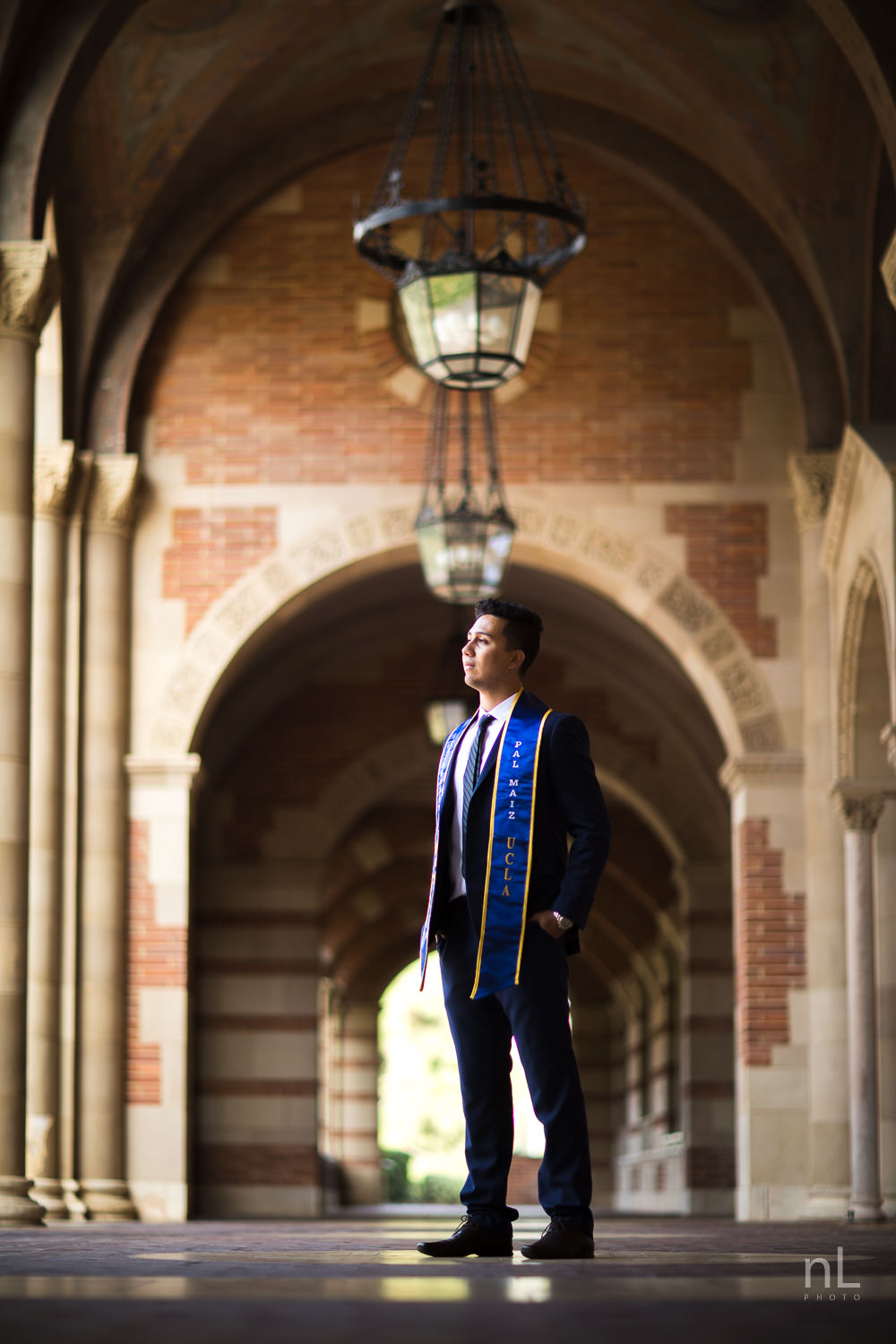 UCLA Class of 2019 graduate wearing suit and tie and graduation sash smiling and standing in Royce Hall arches.