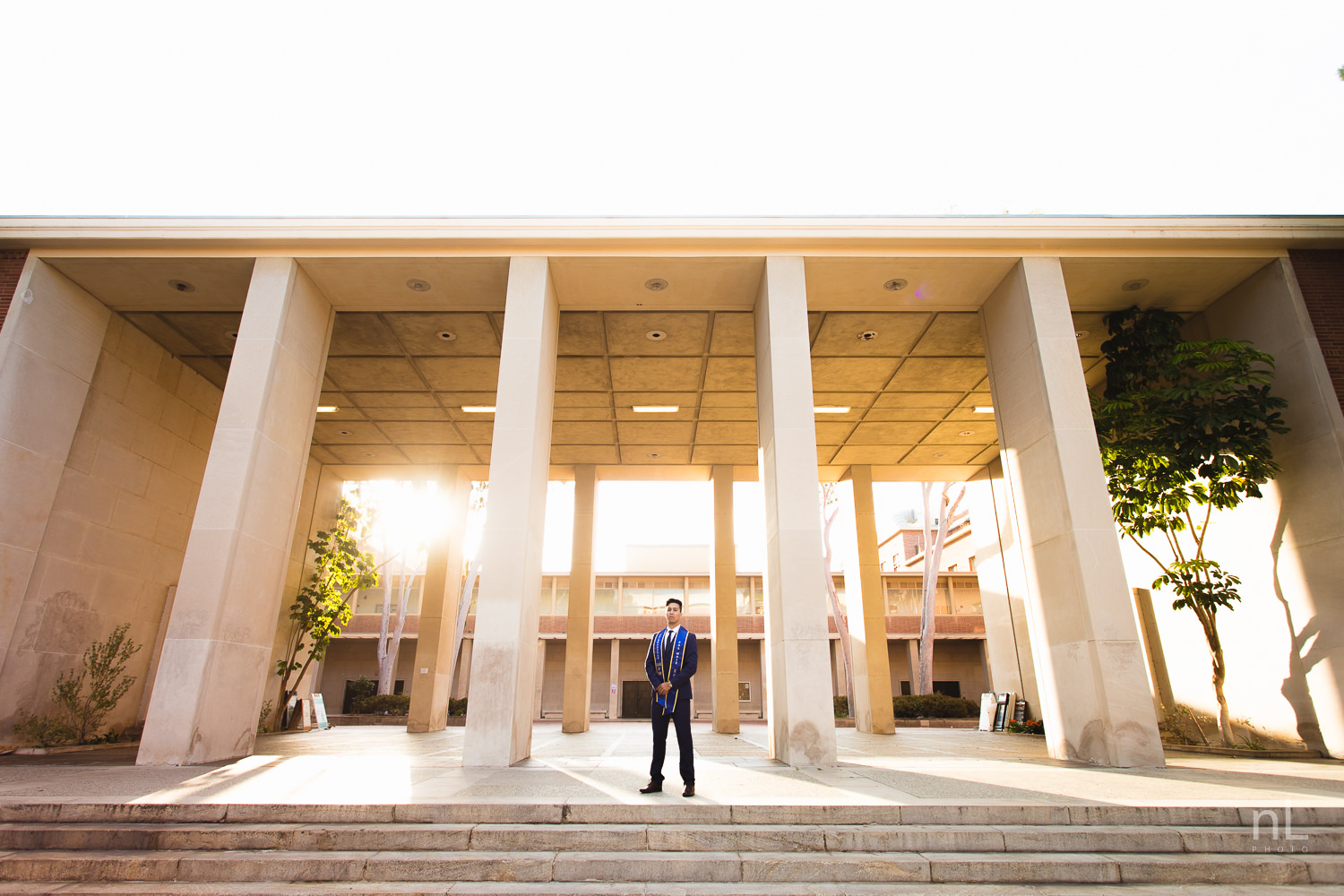 UCLA Class of 2019 graduate wearing suit and tie and graduation sash looking serious in front of Young Hall.