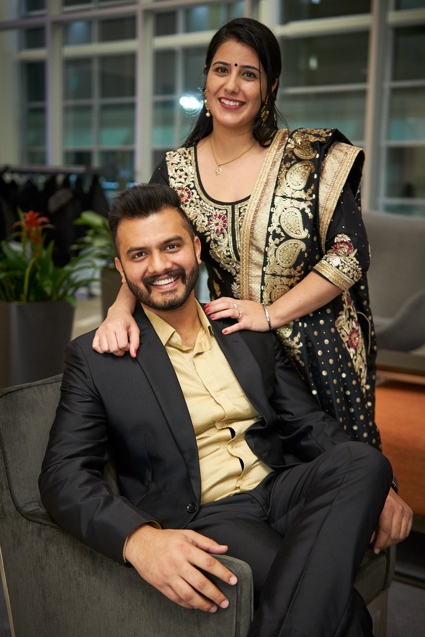 Happy couple posing together at the Calgary Airport Marriott Hotel's annual Christmas party – a sharp black suit and an ornate black and gold lehenga. Elegant portrait by Stance Creative event photography.