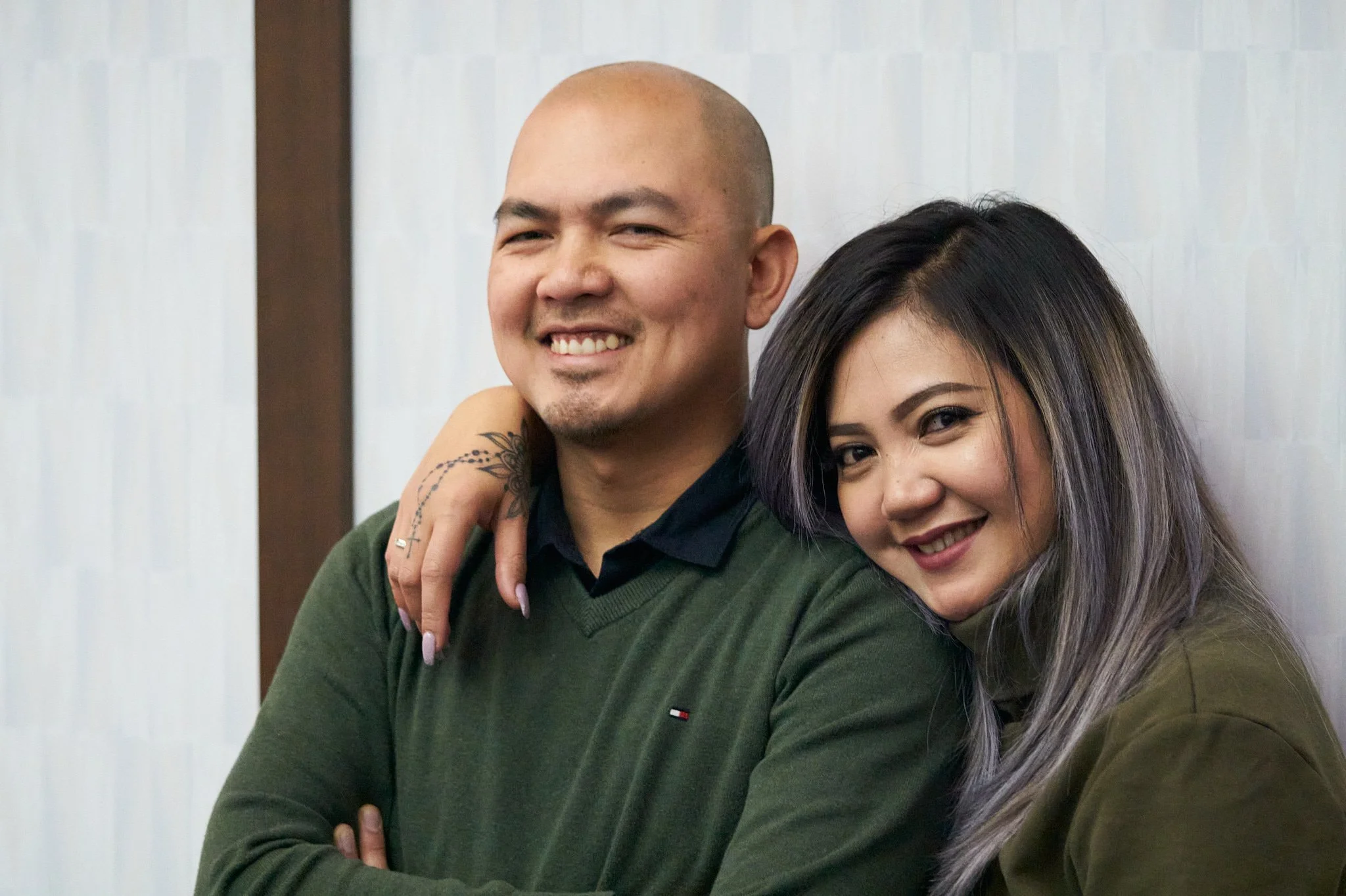 A couple posing arm-in-arm at the Calgary Airport Marriott's annual Christmas Party. Relaxed smiles against a neutral wall. Modern lifestyle portrait by Stance Creative.