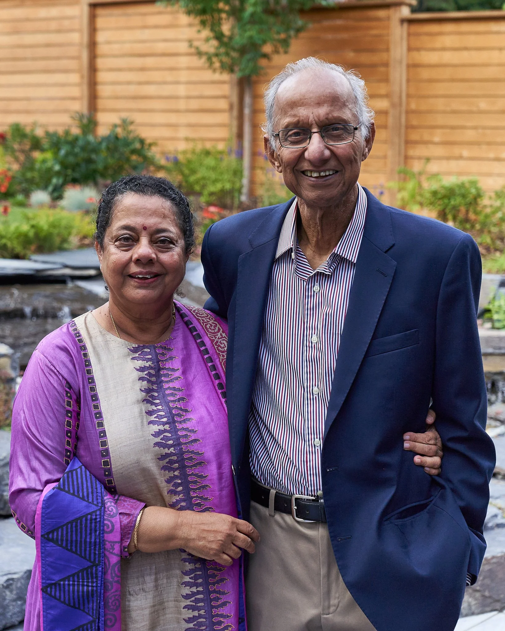 A happy couple standing arm-in-arm in a Calgary garden – her in purple patterned sari, him in navy blazer – radiating warmth and long-lasting love. Classic portrait by Stance Creative.