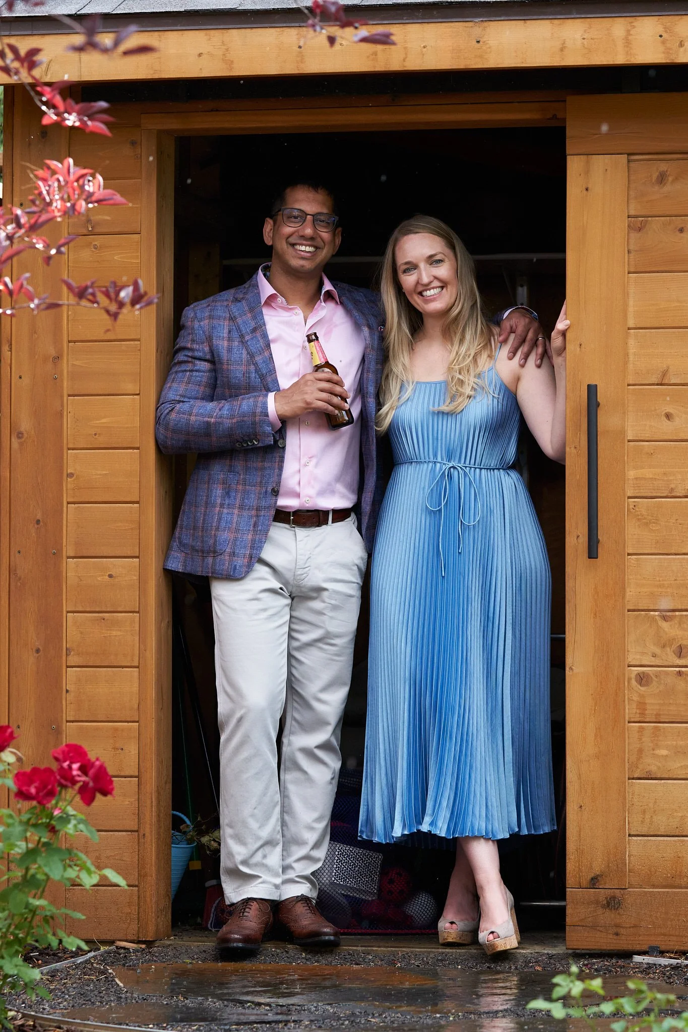 A couple laughing together in a backyard shed doorway – him in plaid blazer with beer, her in blue pleated dress – relaxed and happy in Calgary. Candid outdoor portrait by Stance Creative.