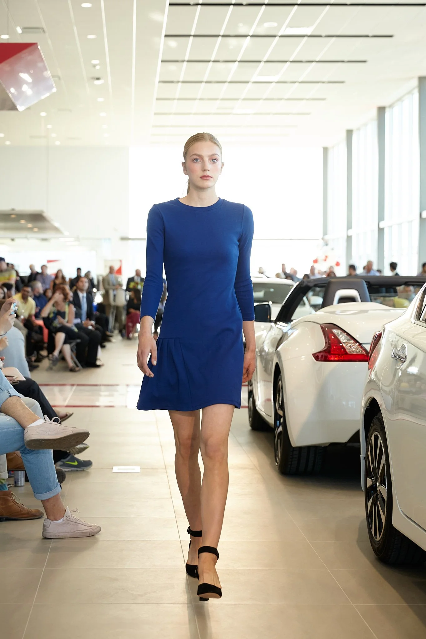 Confident model walking the runway in a sleek blue long-sleeve dress amid luxury Nissan cars and attentive audience. High-fashion moment by Stance Creative Calgary event photographer.